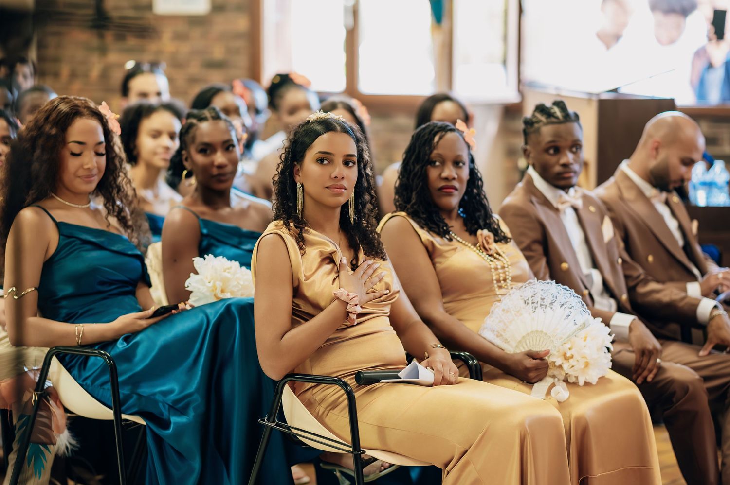 Bridesmaids in gold and teal satin dresses seated at wedding ceremony holding fans.
