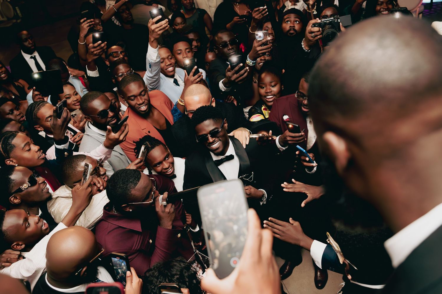 Crowd of wedding guests taking selfies and photos with groom at a lively African wedding reception.