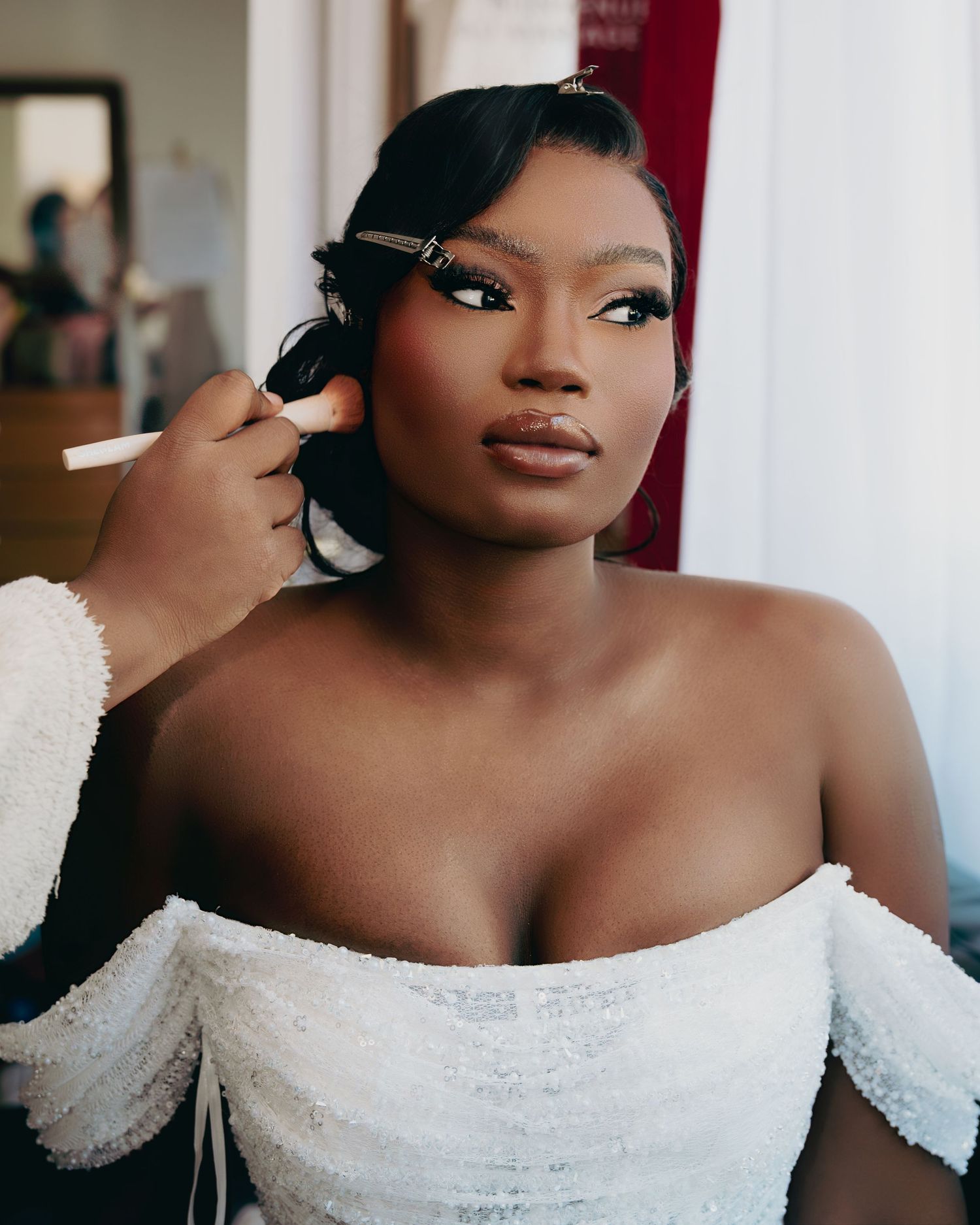 Beautiful dark-skinned bride in off-shoulder white gown gets makeup applied by a professional artist.