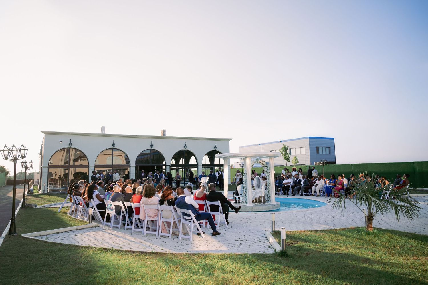 Outdoor wedding ceremony by a pool at a white venue with arched windows at dusk.