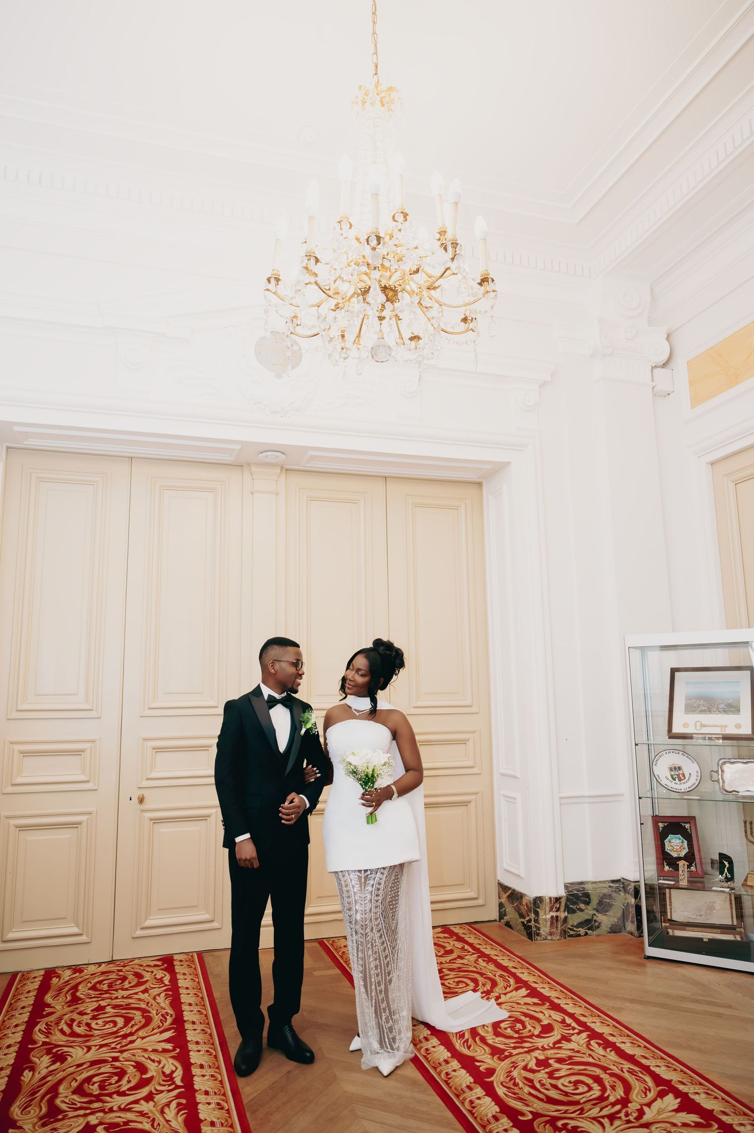 Bride and groom standing in an elegant white ballroom with crystal chandelier and red patterned carpet.