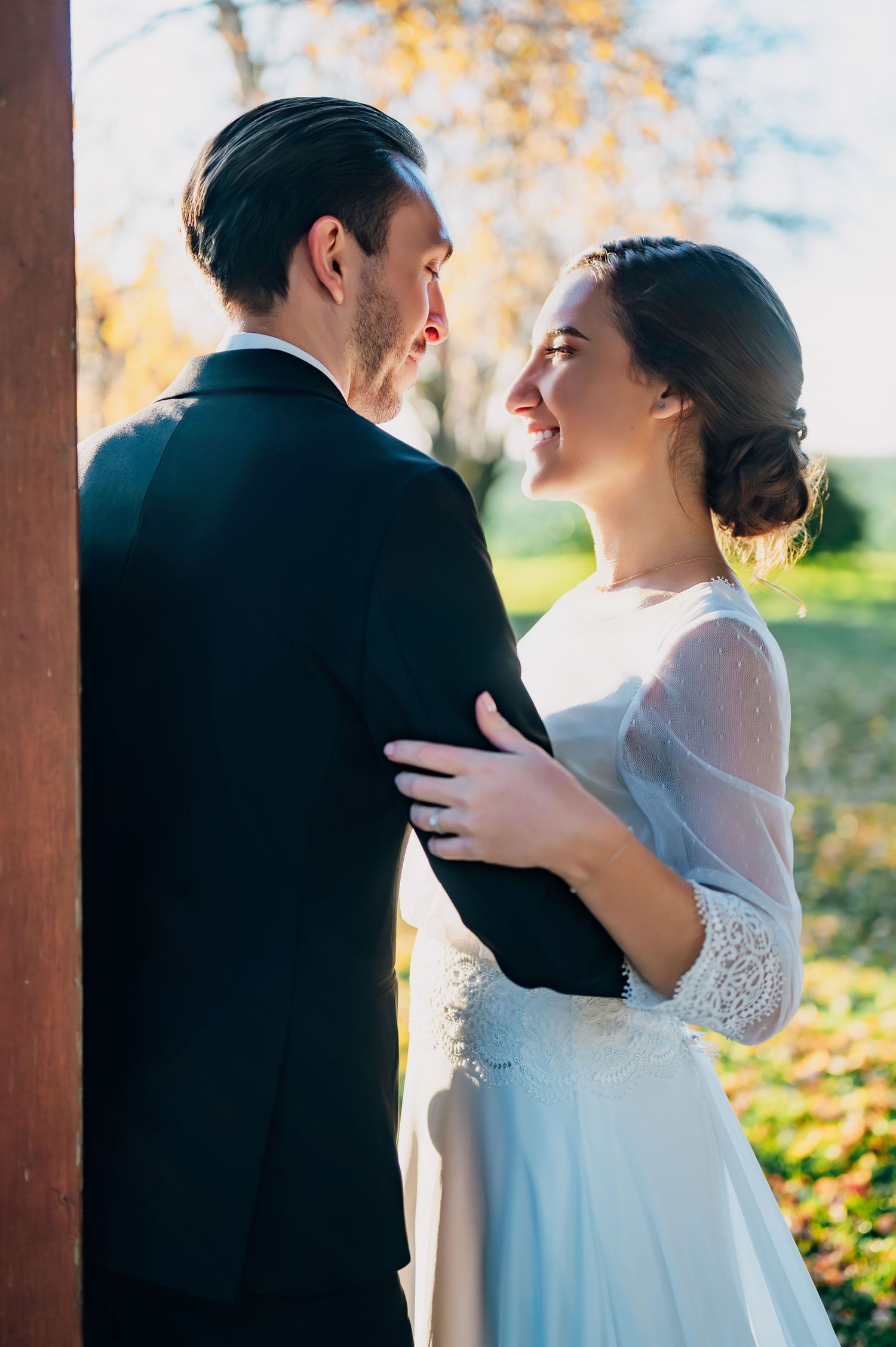Bride in sheer-sleeve white gown smiling at groom in black suit during golden hour outdoor wedding portrait.