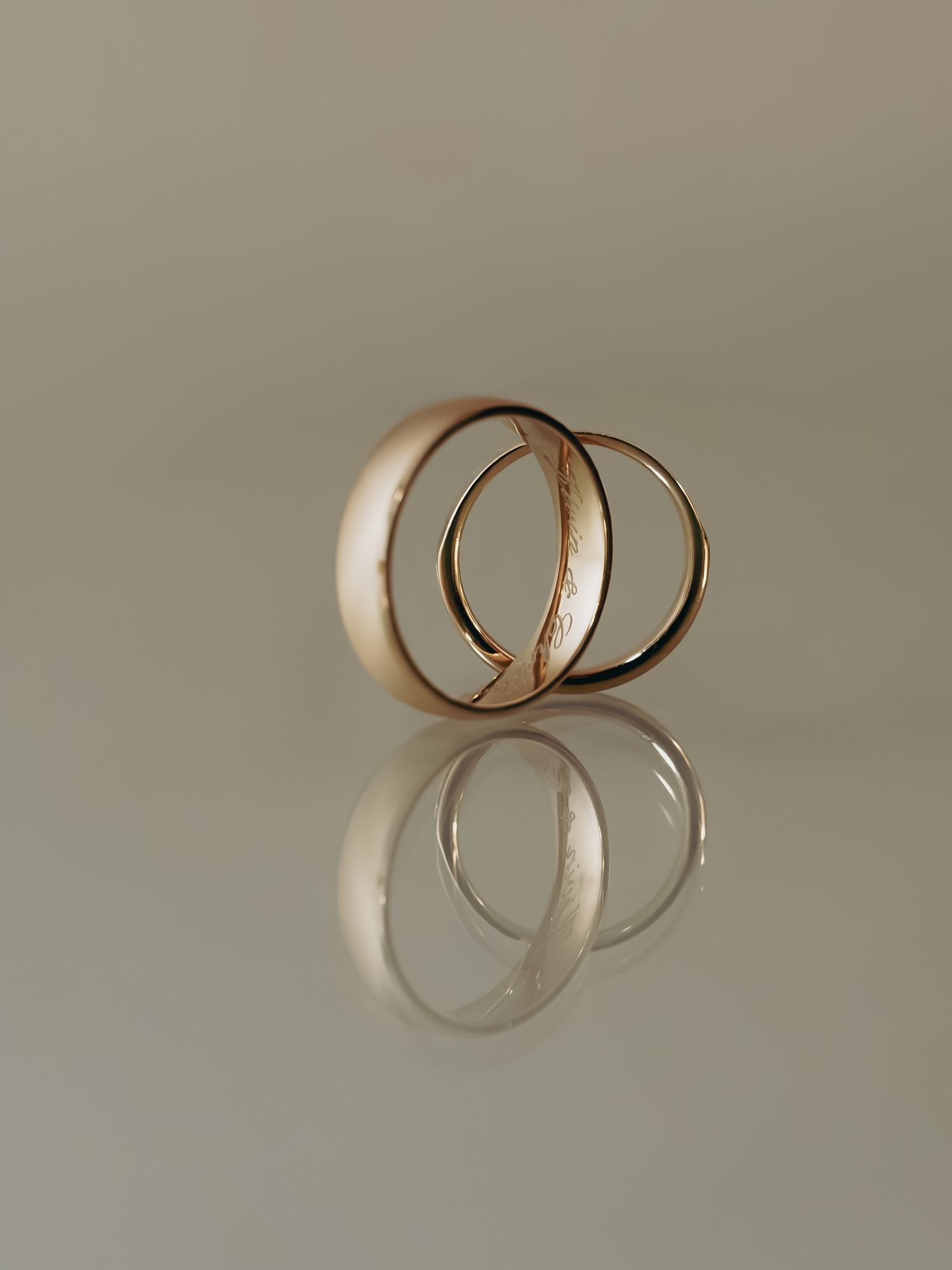 Two interlocked gold wedding bands reflected on a smooth neutral surface in a minimalist composition.