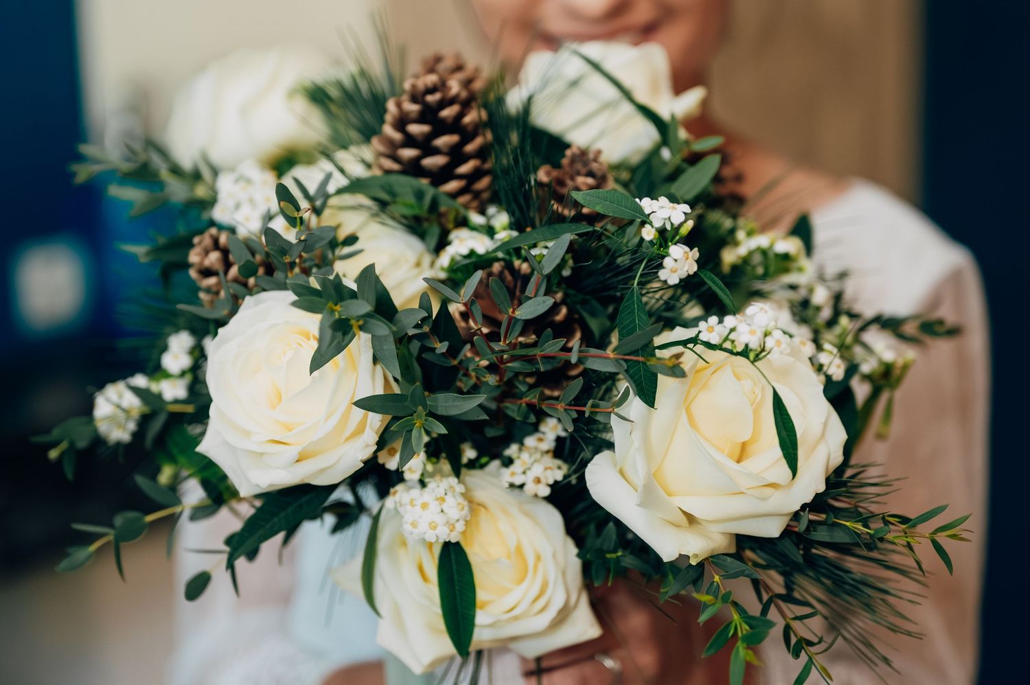 Close-up of winter wedding bouquet featuring white roses, pine cones, and greenery held by bride.