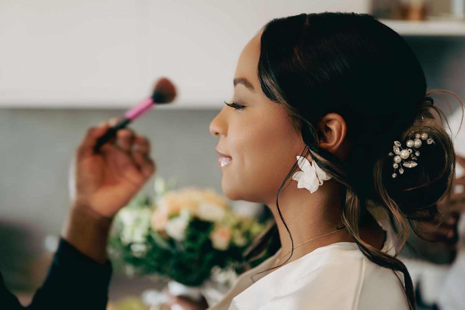 Bride gets blush applied with a makeup brush while wearing floral earrings and hair accessory.