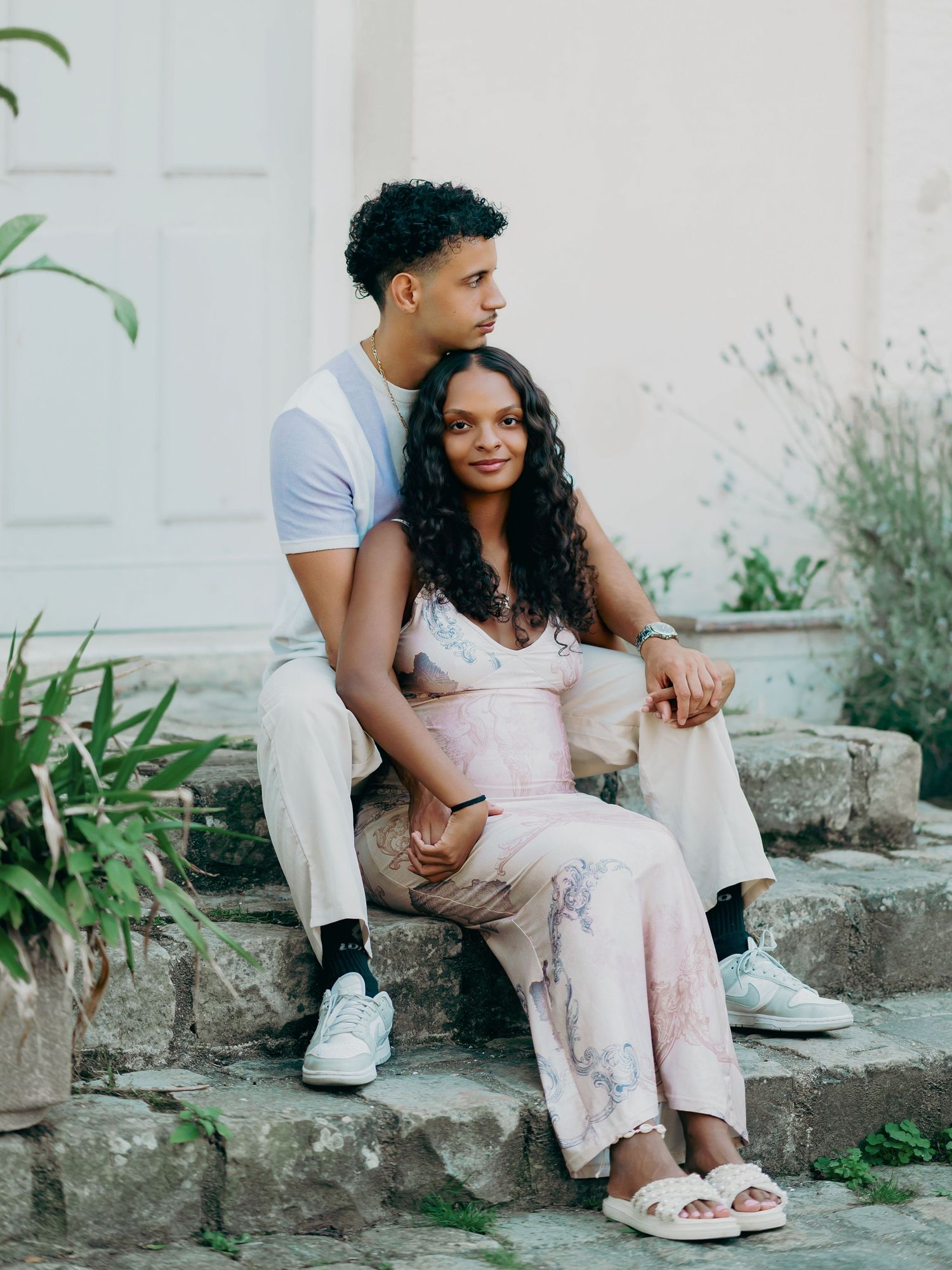 Couple sitting on stone steps outside a white building, woman in floral maternity dress smiling softly.