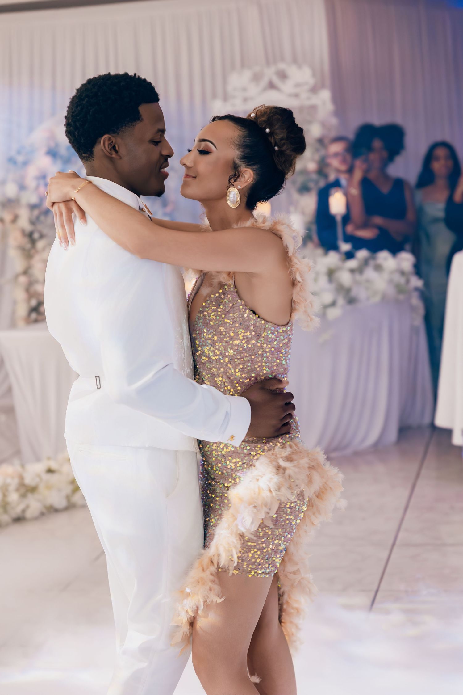 Bride in gold sequin mini dress with feathers shares romantic first dance with groom in white suit.