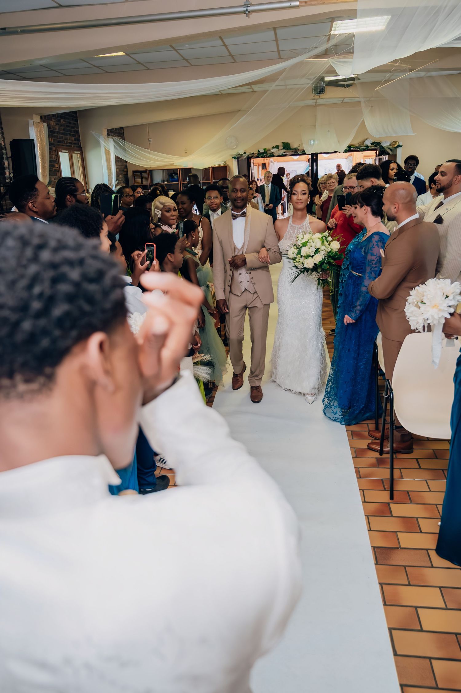 Bride and escort walk down wedding aisle as guests photograph the ceremony procession.