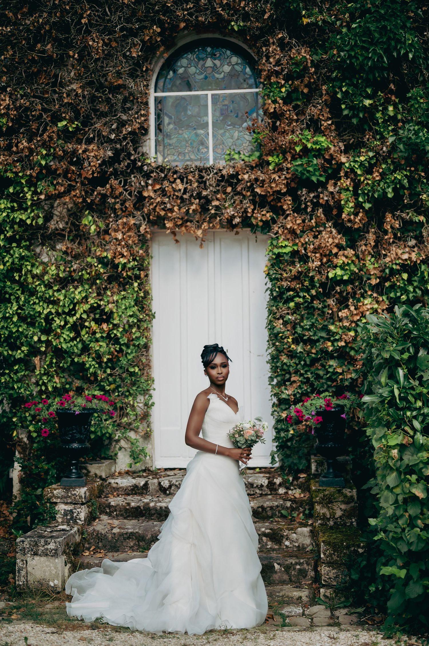 Elegant bride in white ruffled gown holds bouquet before ivy-covered stone wall with arched white door.