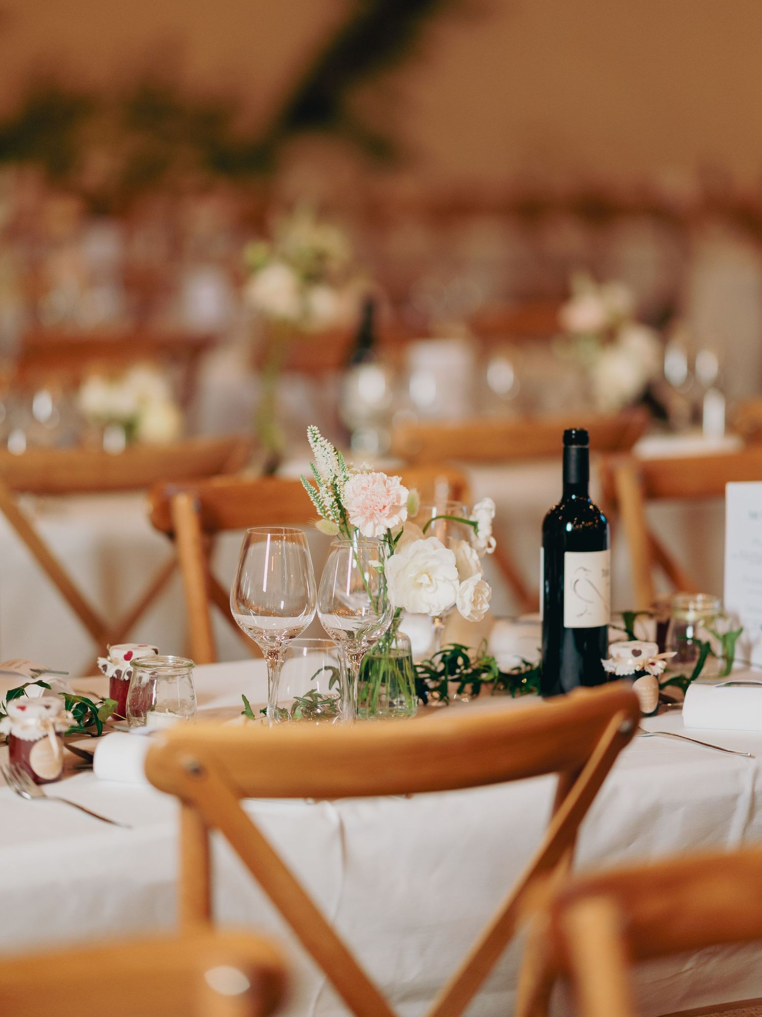 Elegant wedding reception tables with white linens, wooden cross-back chairs, floral centerpieces and wine.