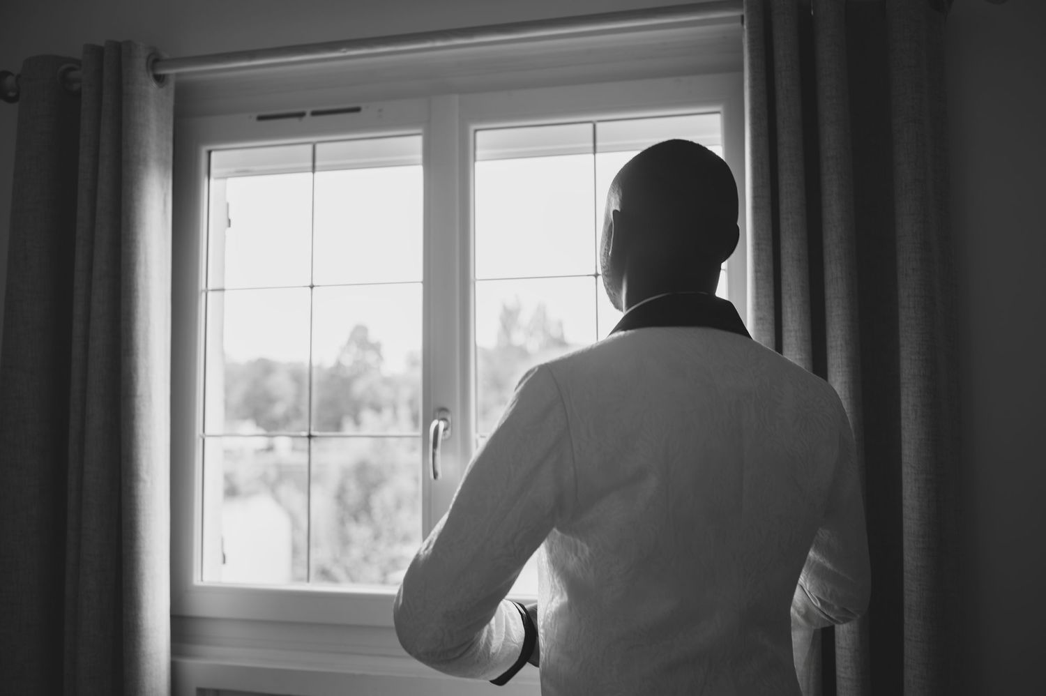 Black and white photo of groom in white shirt gazing out a bright window.