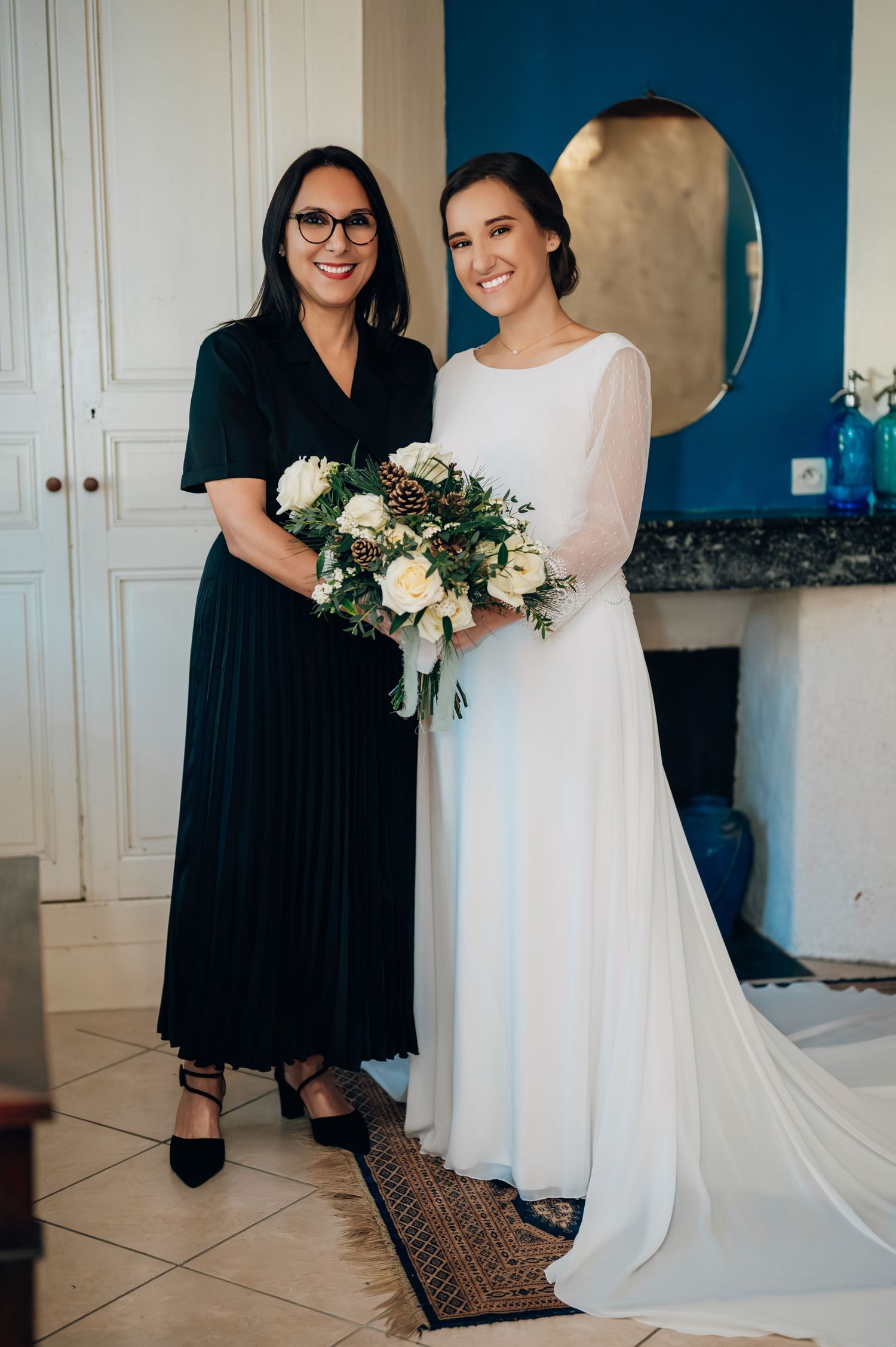 Bride in white gown holding winter bouquet with roses and pine cones stands with woman in black dress indoors.