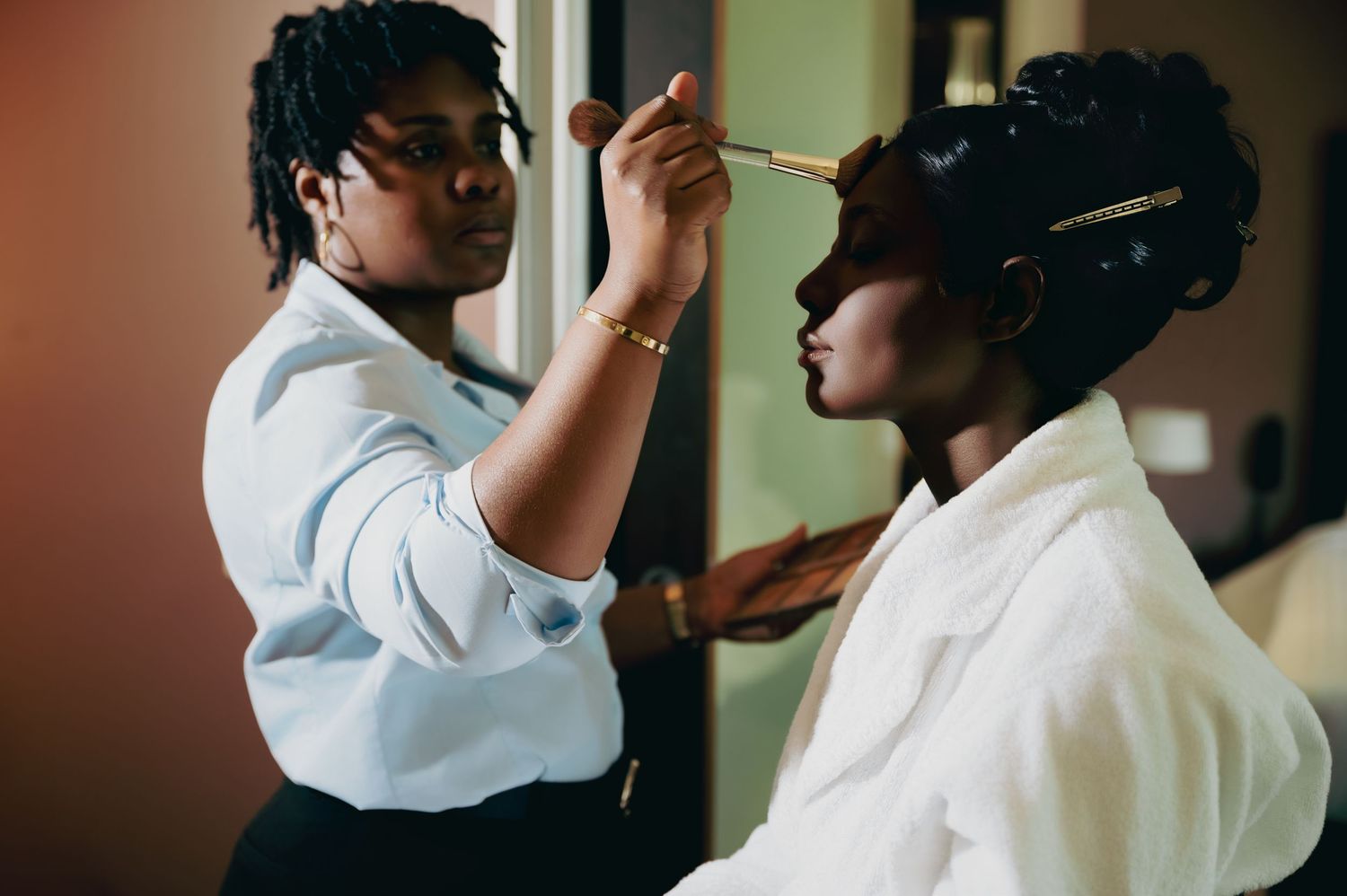A makeup artist applies eyeshadow to a bride during wedding preparation in a softly lit room.