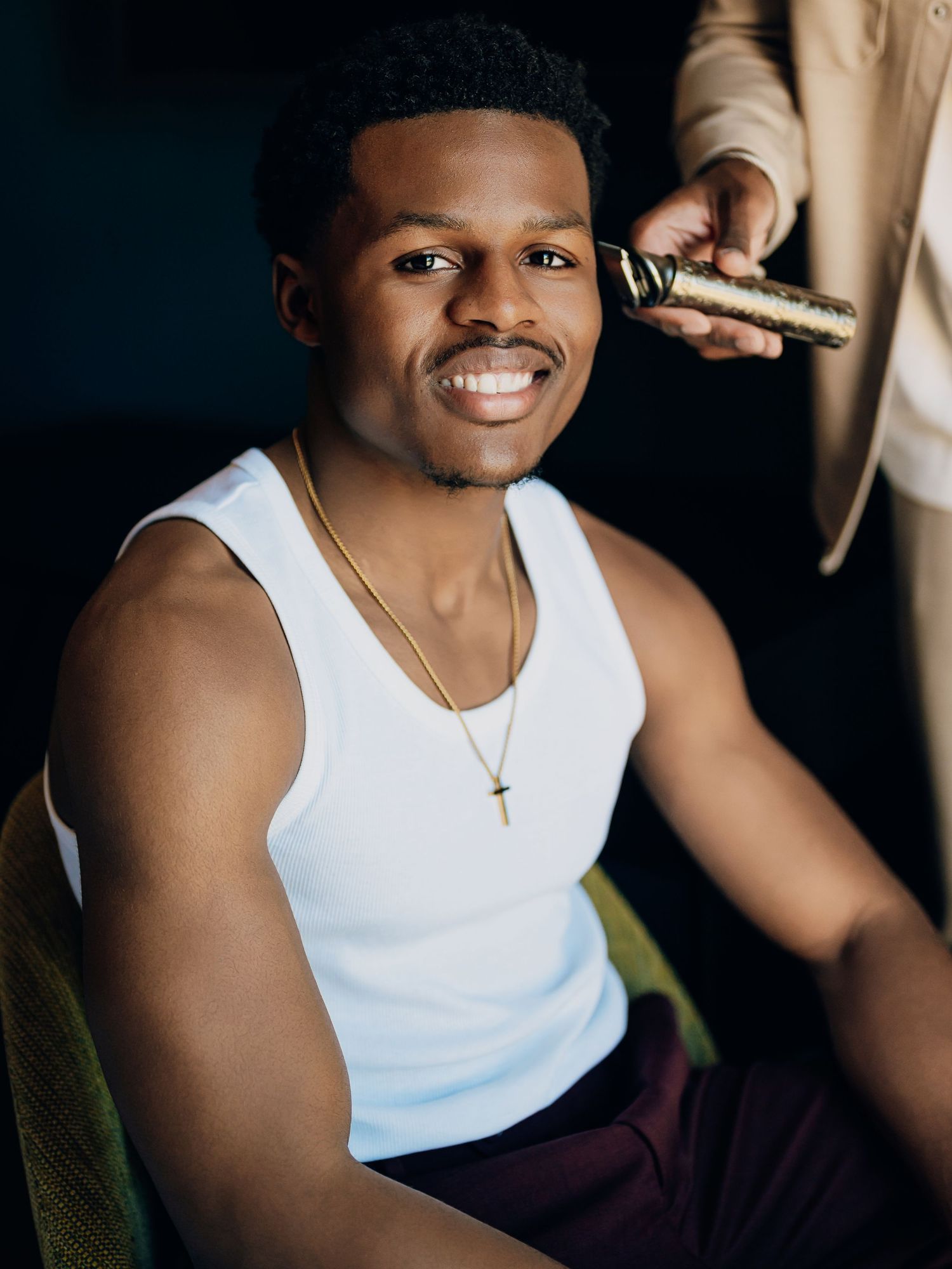 Smiling groom getting a haircut before his wedding day, wearing a white tank top and gold cross necklace.