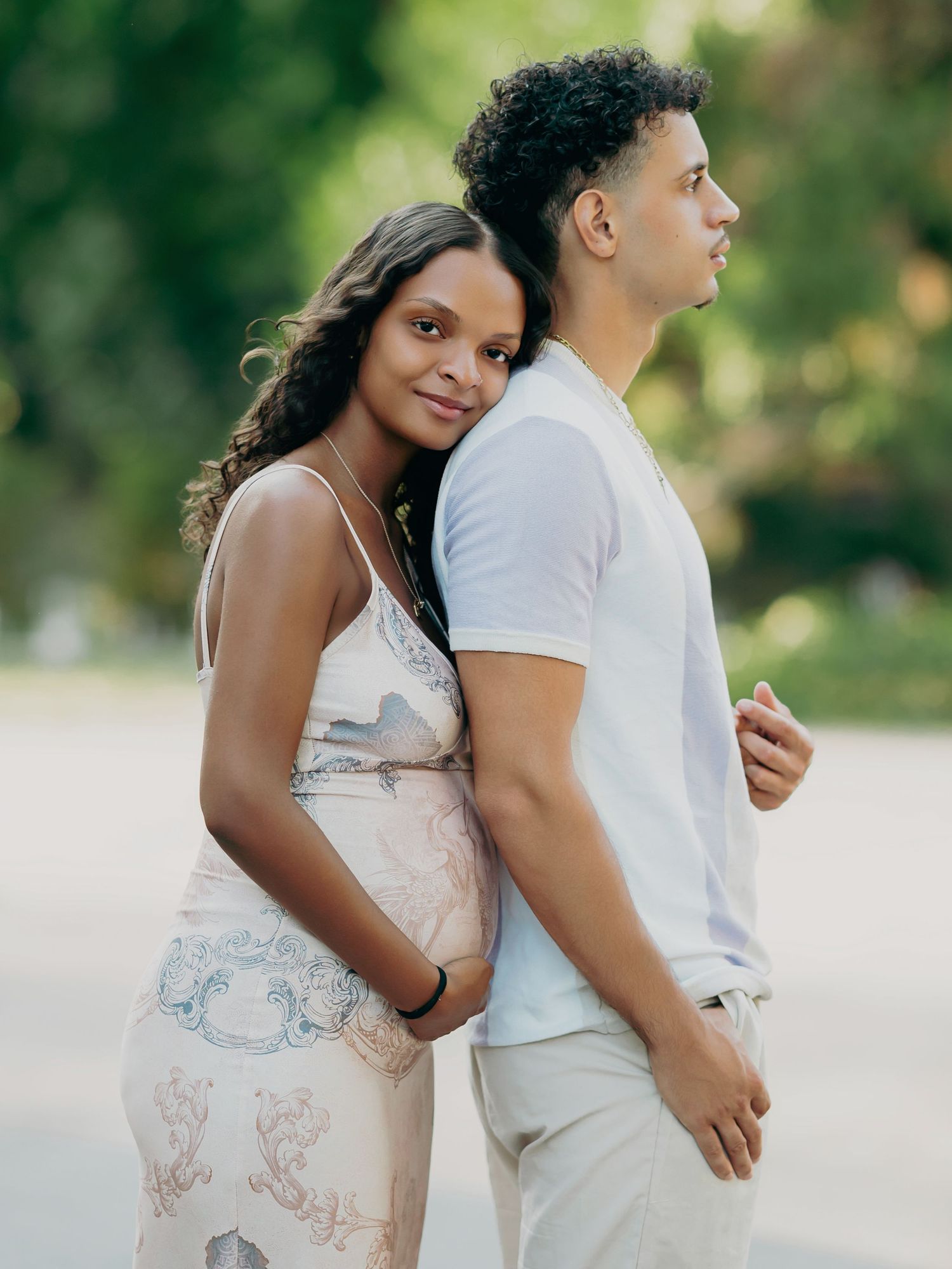 Expectant couple leans back to back in soft natural light, woman in elegant floral maternity dress.