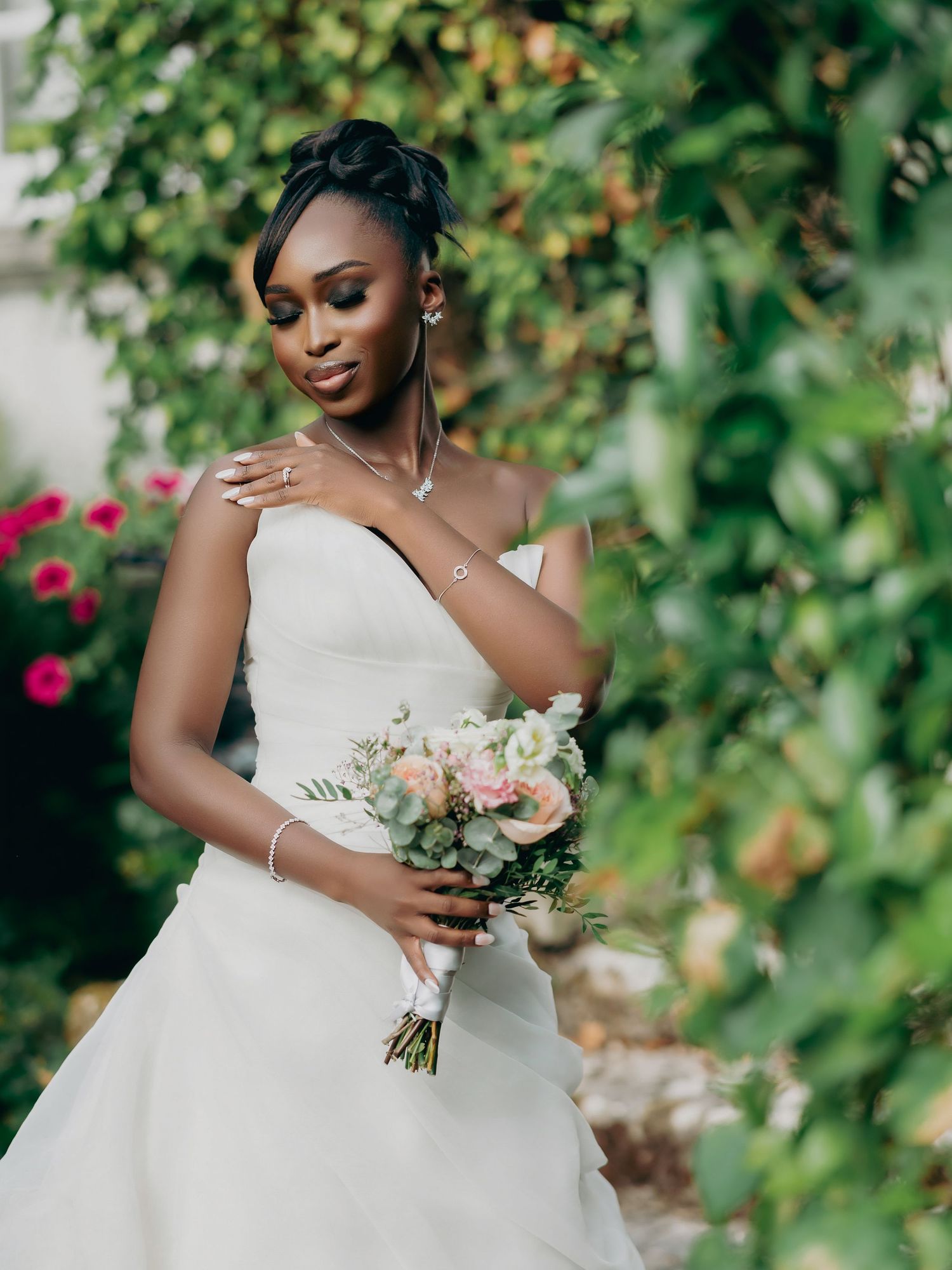 Beautiful Black bride in a white wedding dress holds a pink and green bouquet among lush greenery.