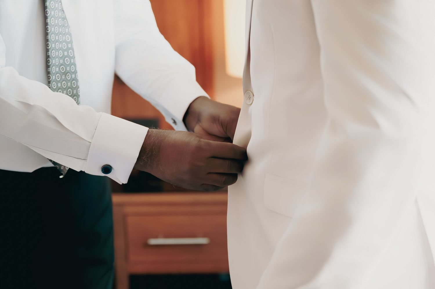 A man helping another adjust his white suit jacket during wedding preparations.
