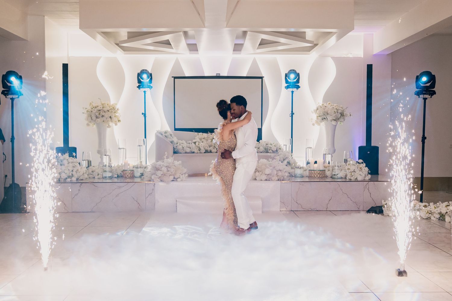 Couple shares first dance on cloud-covered floor with fireworks and white floral decor at wedding.