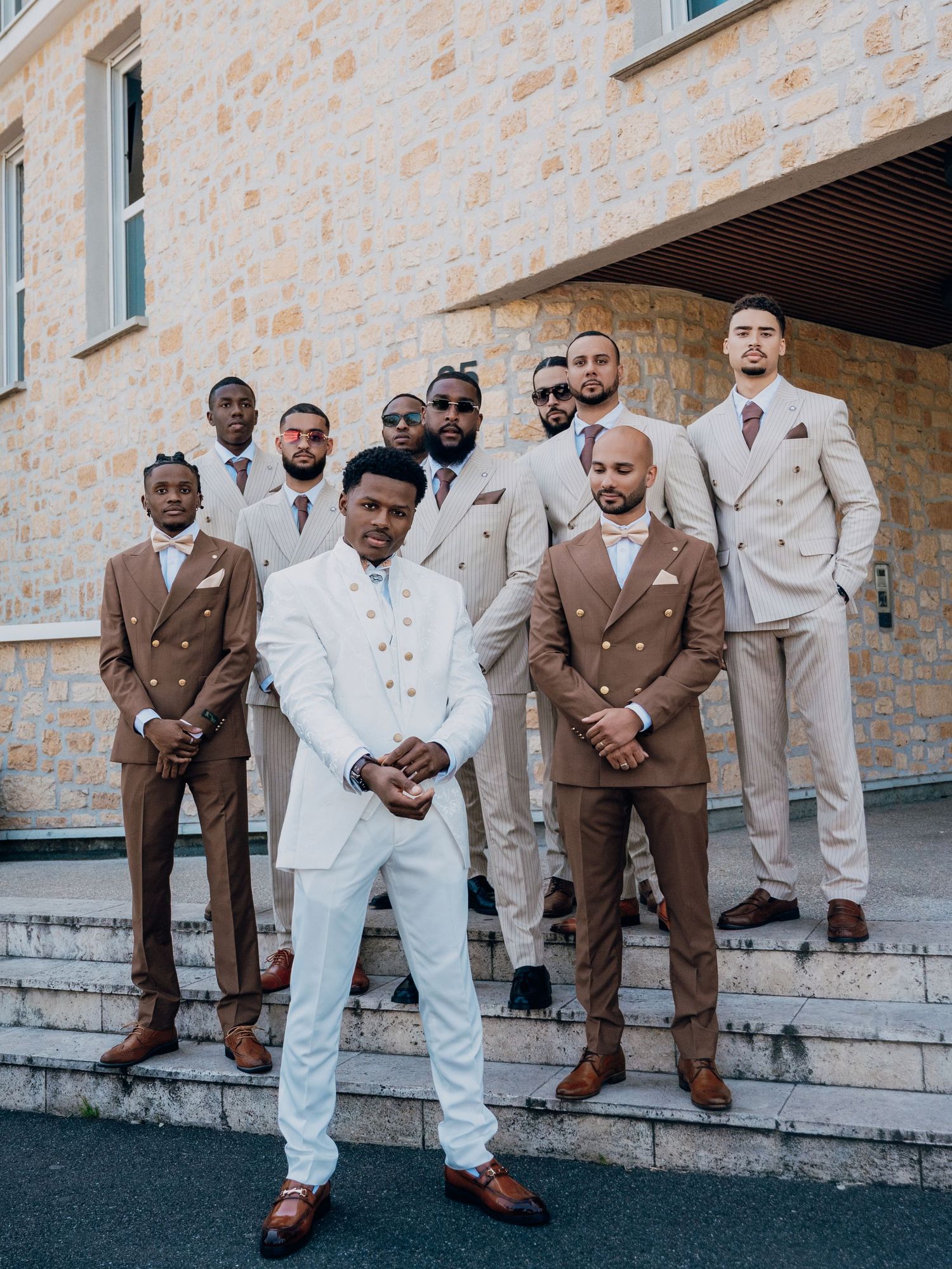 Groomsmen in brown and beige double-breasted suits with groom in white suit posing on steps.