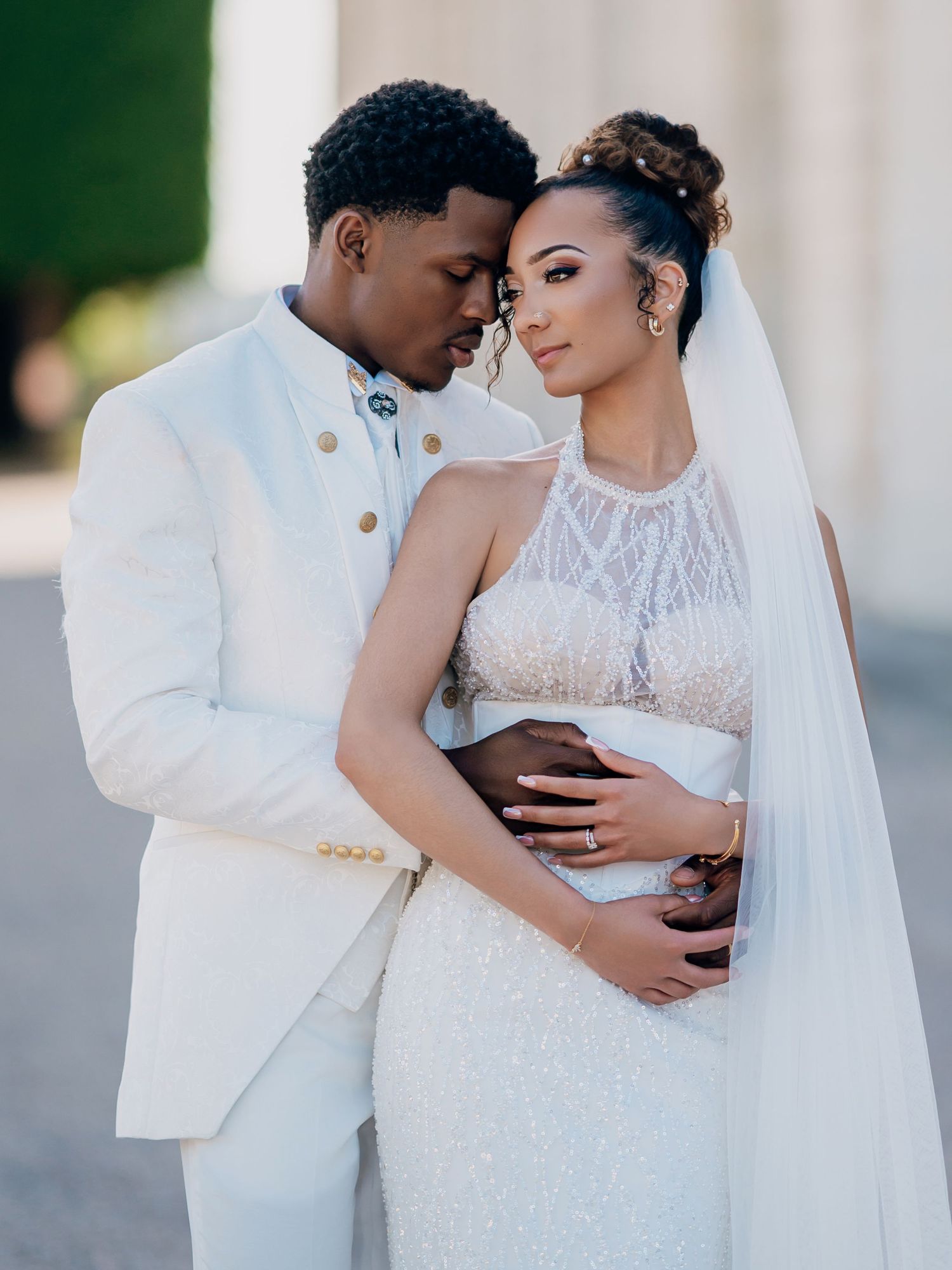 A Black couple in wedding attire embrace tenderly, groom in white suit, bride in beaded gown with veil.