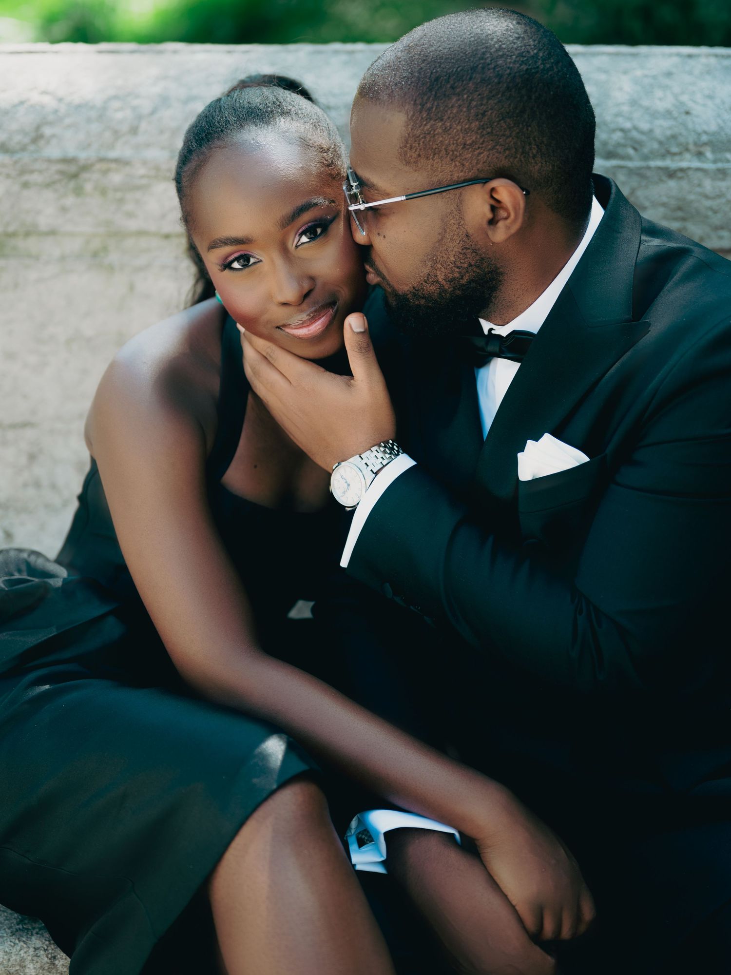 Tender Black couple in formal black outfits share a sweet cheek kiss on stone steps.