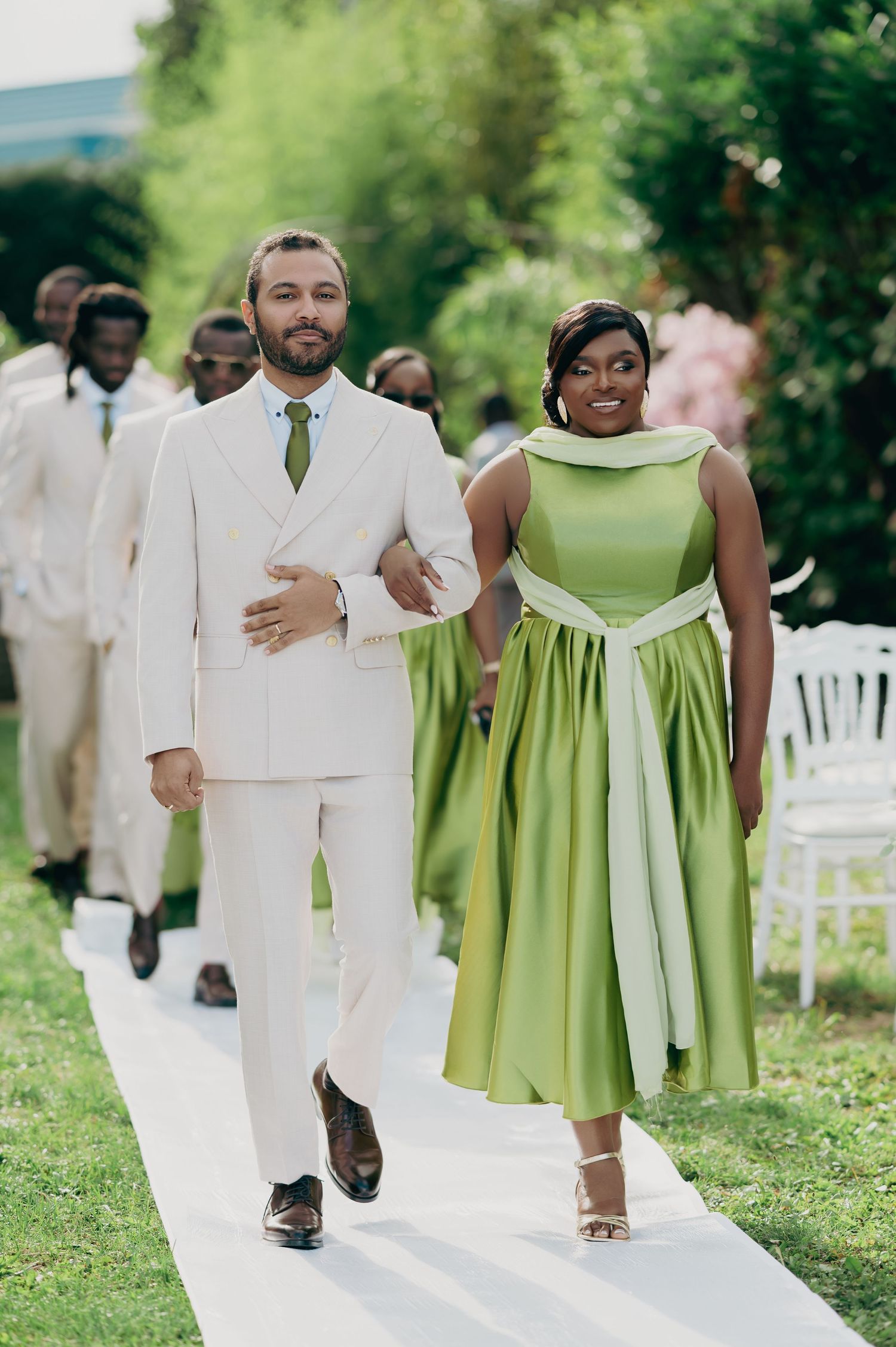 A groomsman in a white suit escorts a bridesmaid in a green midi dress down the outdoor wedding aisle.