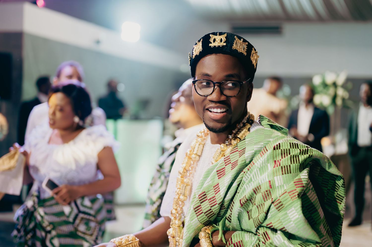 Smiling groom in green kente cloth traditional African attire and black crown at wedding reception.