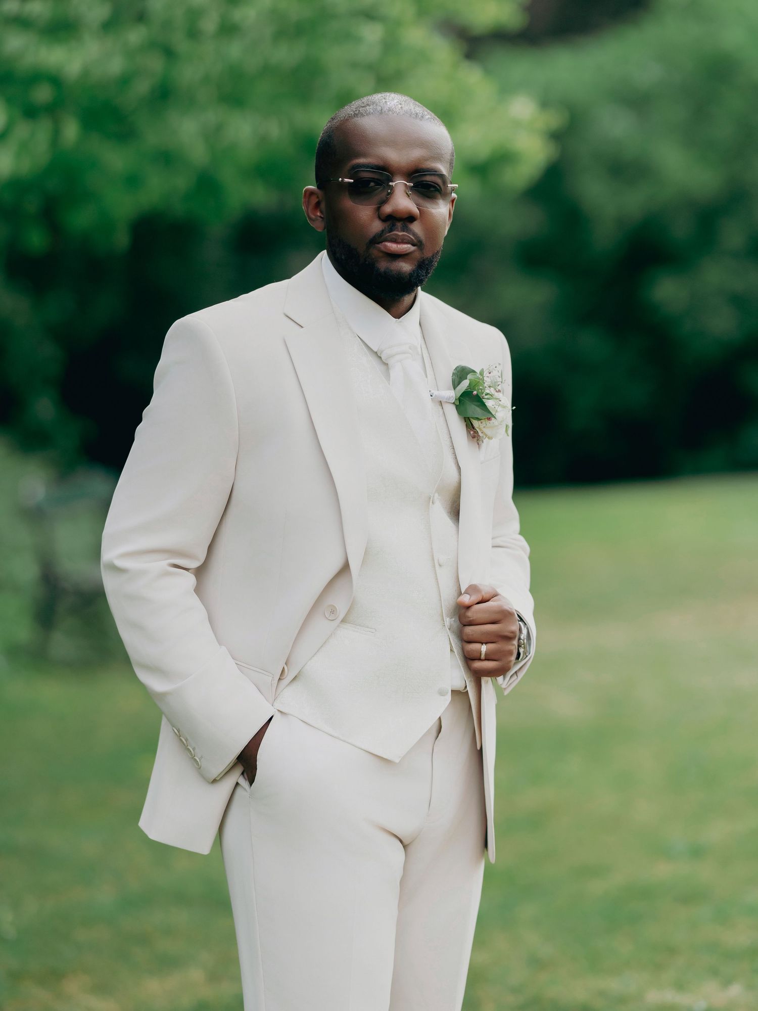 Dapper Black groom in a beige three-piece suit with floral boutonniere poses in a garden setting.