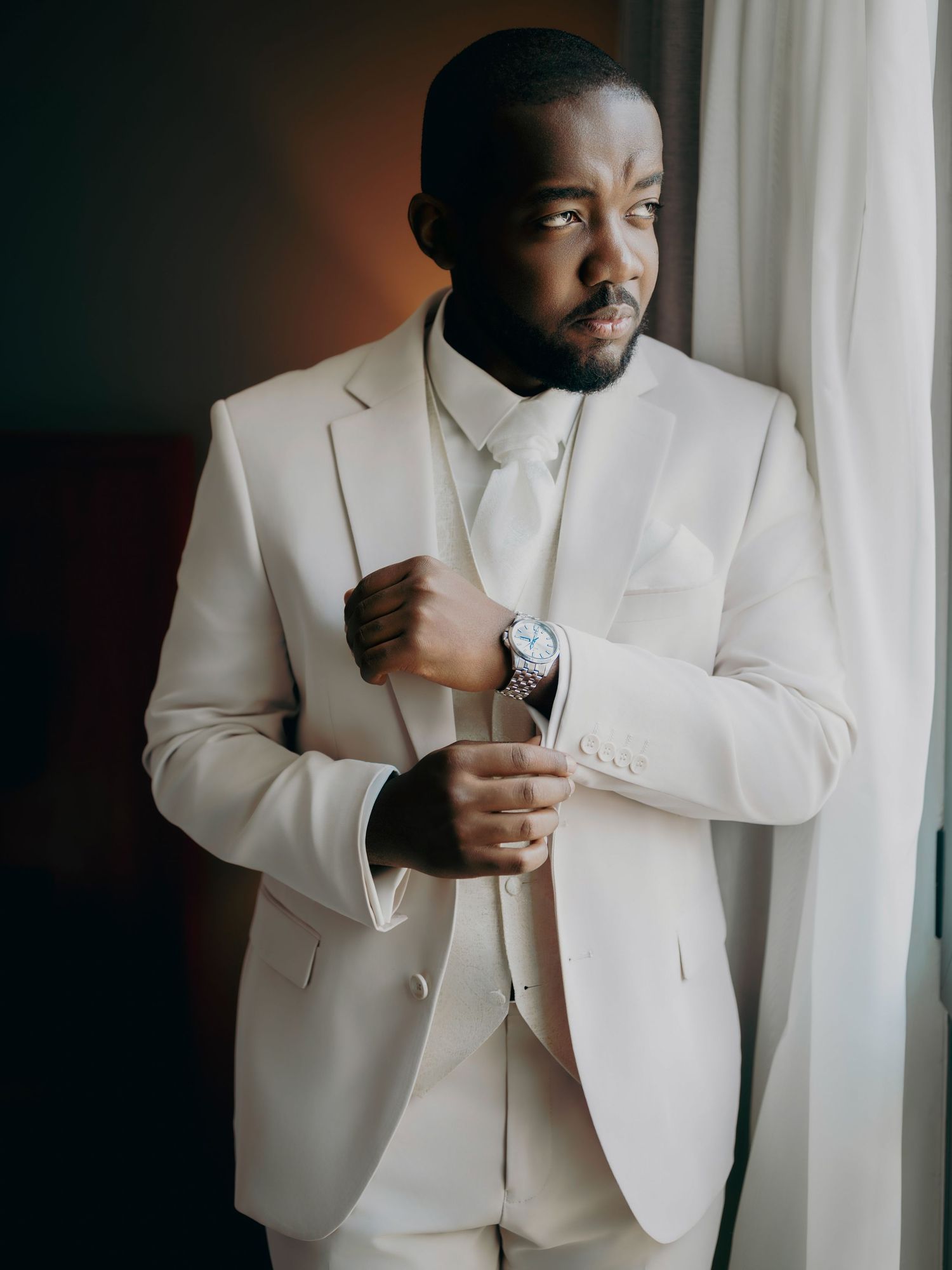 Elegant groom in a cream suit adjusting his cufflinks by a window.