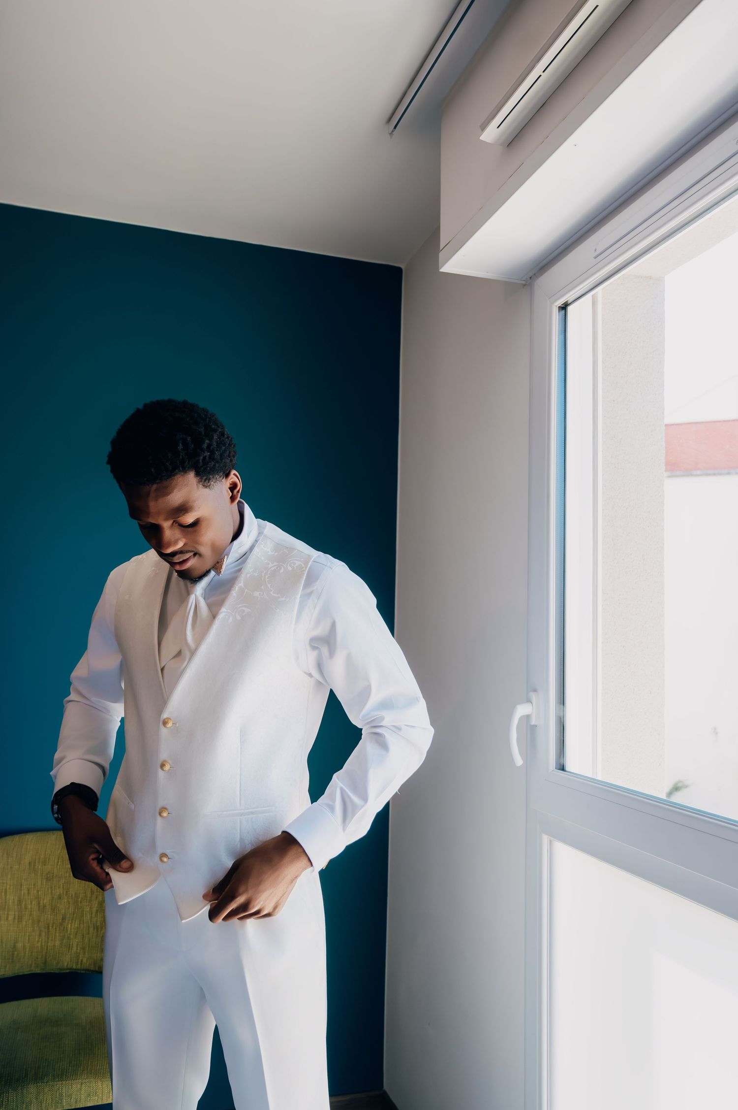 Dapper groom in all-white suit and vest adjusting his outfit near a window before the wedding ceremony.