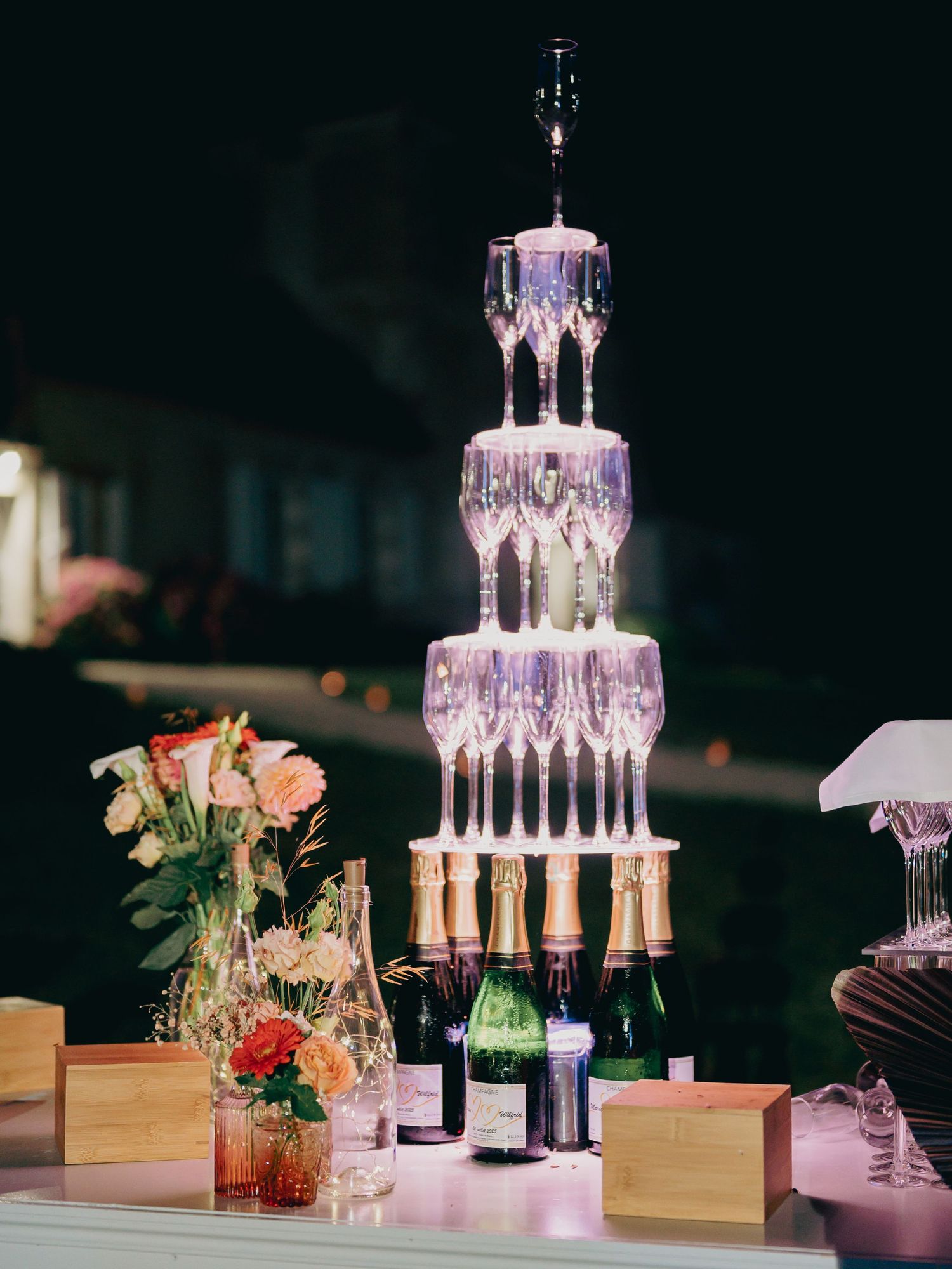 Illuminated champagne tower display with bottles and floral arrangements at elegant wedding reception.
