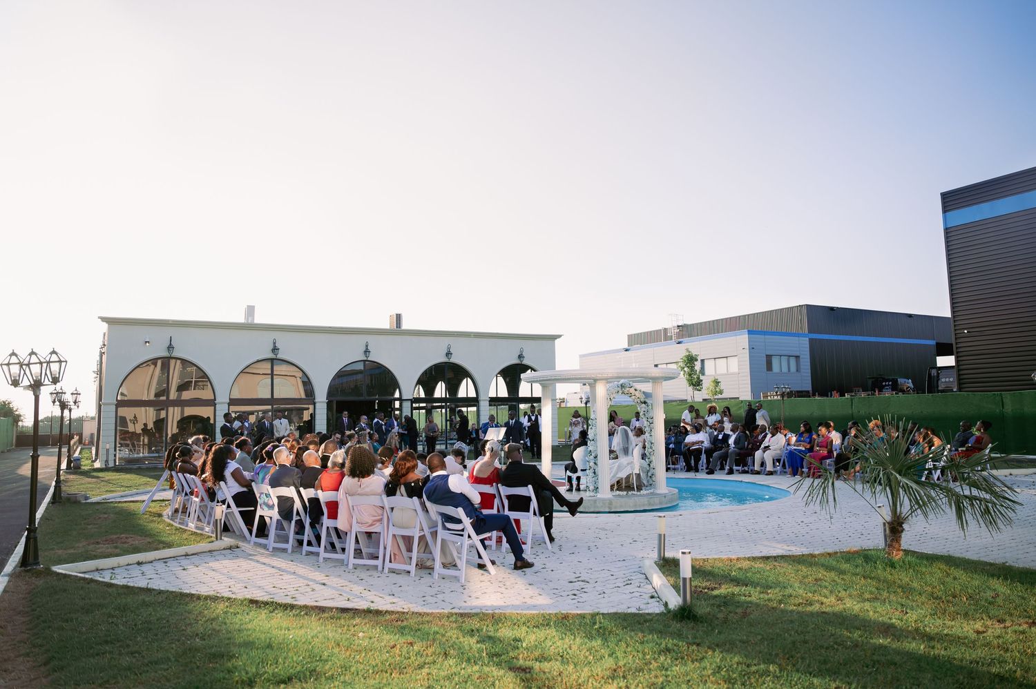 Outdoor wedding ceremony beside a pool at dusk with guests seated before a white gazebo altar.