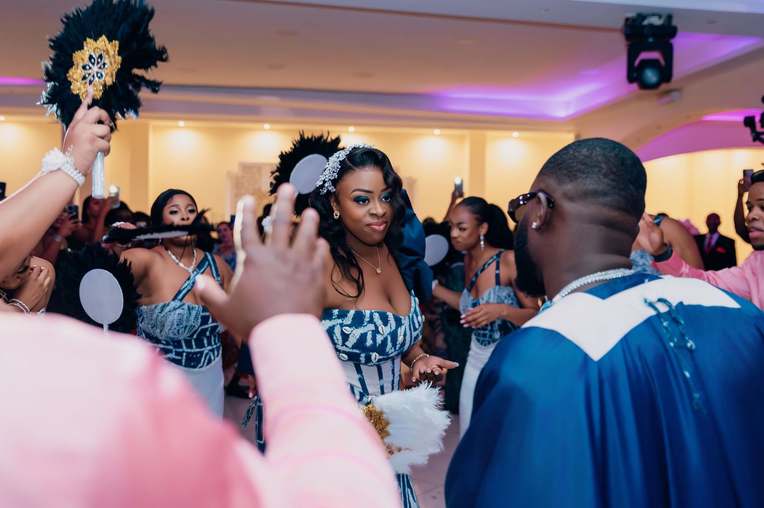 Bride in blue and white dress surrounded by celebrating guests waving decorative fans at reception.