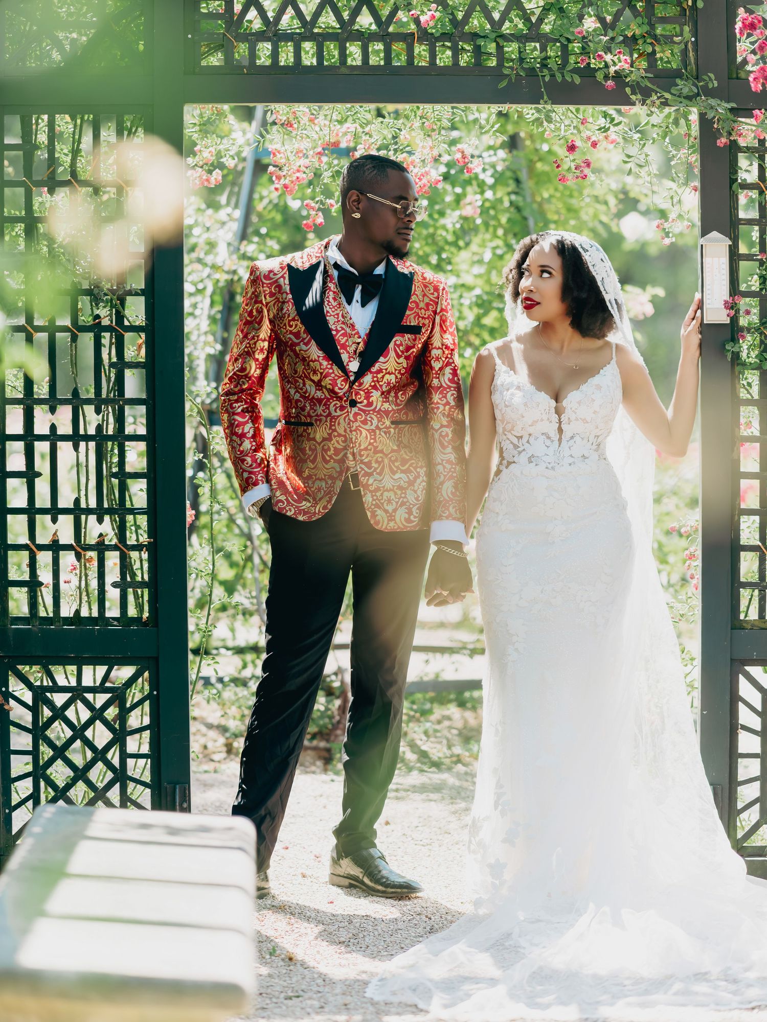 Bride and groom stand in sunlit garden gazebo, groom wearing ornate red blazer and bride in lace dress.