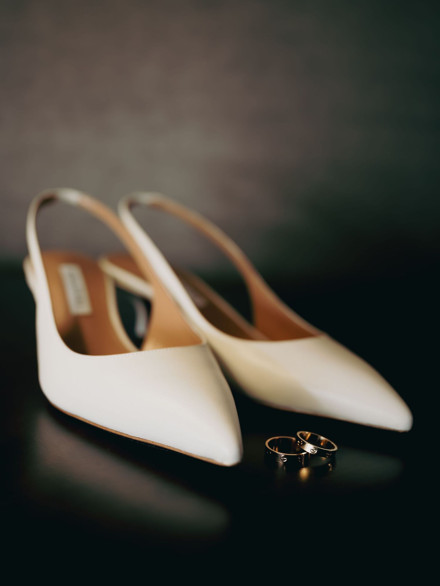 White slingback pointed-toe bridal heels with gold wedding rings placed in front on dark surface.