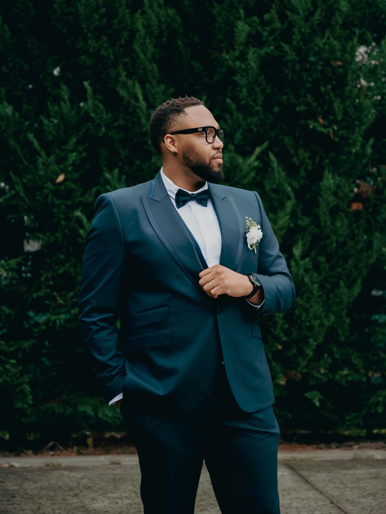 Dapper groom in navy tuxedo with boutonniere posing outdoors before wedding.