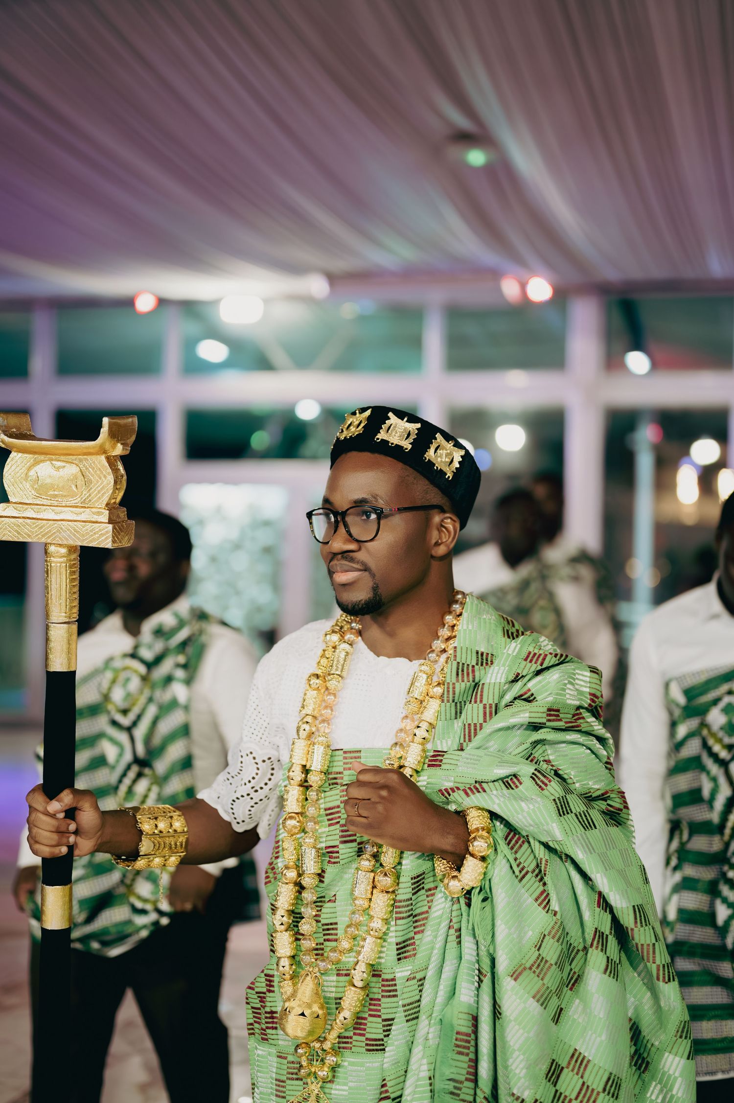 Groom in traditional Ghanaian kente cloth and gold jewelry holding ceremonial staff at wedding.