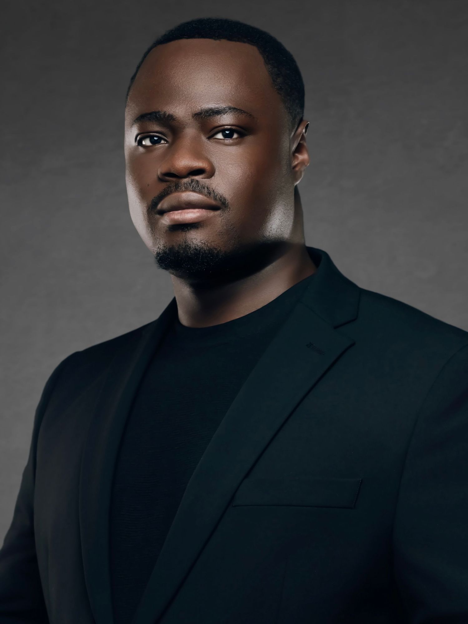 A confident Black man in a black blazer and turtleneck poses in a professional studio portrait.