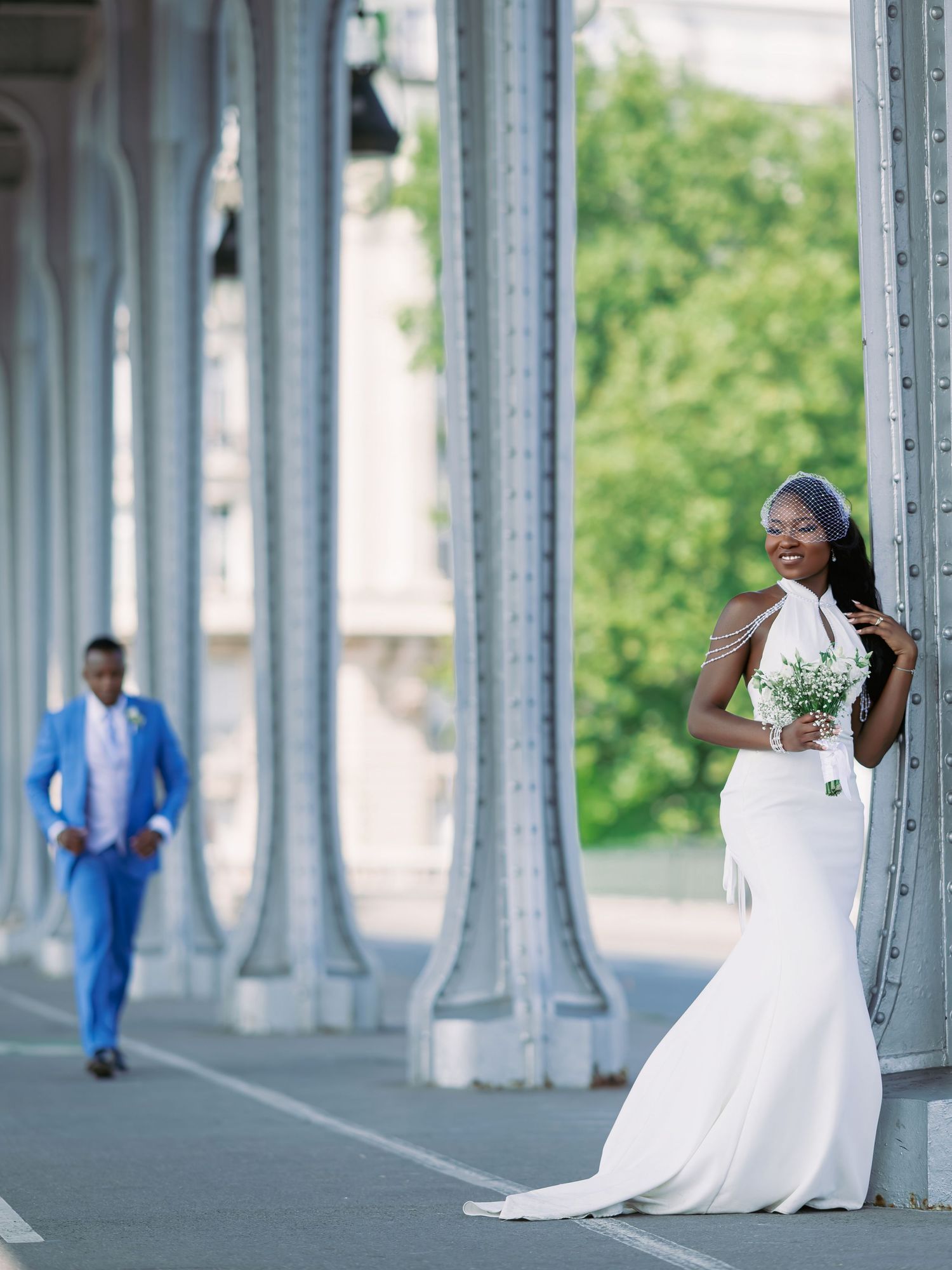 Elegant Black bride in white halter mermaid gown holds bouquet by stone columns as groom stands in blue suit.