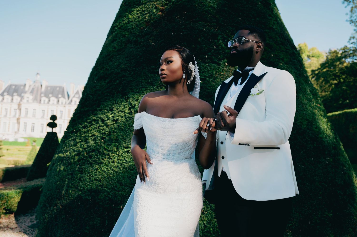 Elegant bride and groom pose before a grand chateau garden with sculpted hedges on their wedding day.