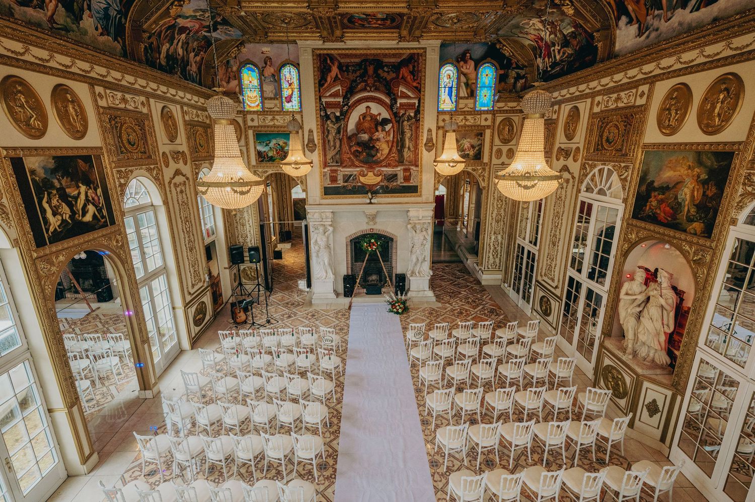 Ornate wedding ceremony hall with frescoed walls, chandeliers, and rows of chairs on a white aisle.