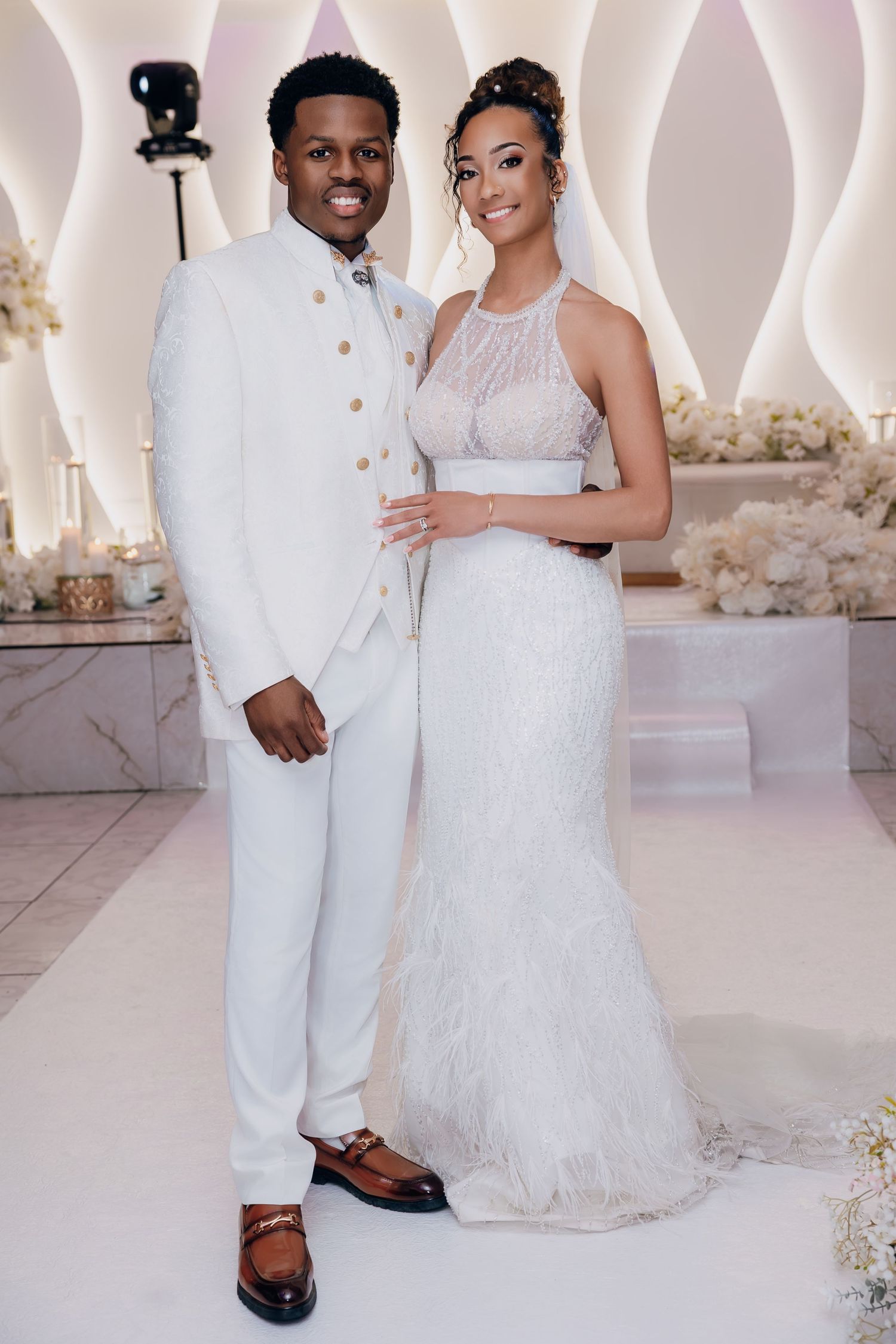 Bride in white feathered halter gown and groom in white suit posing at wedding.