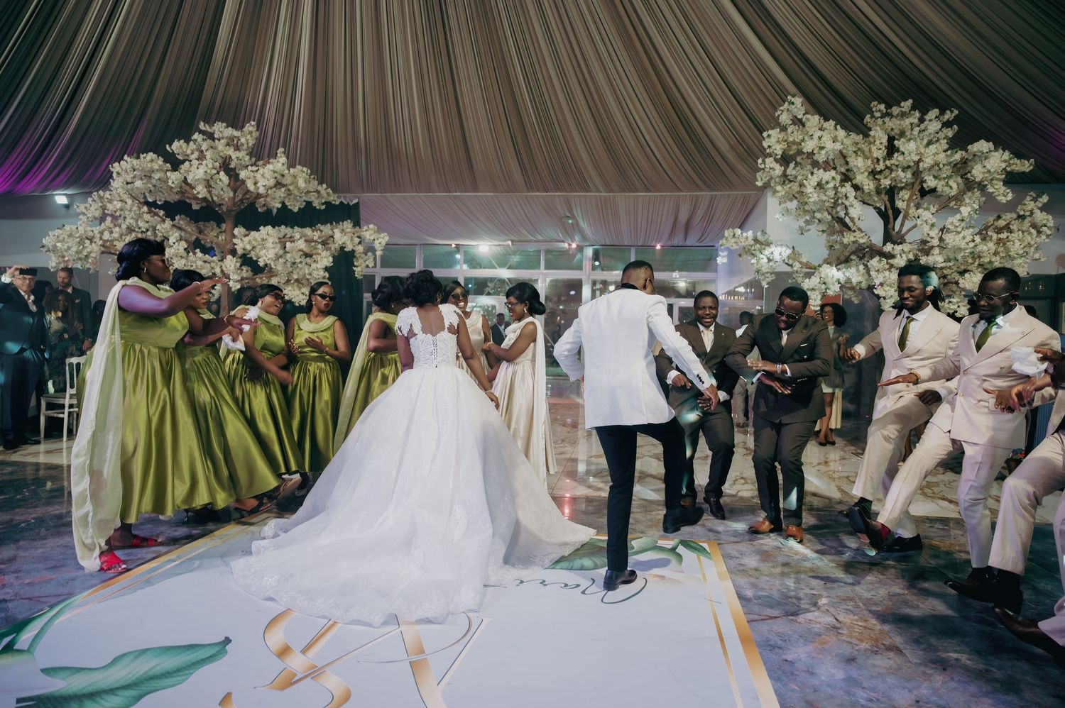 Bride and groom celebrate with wedding party in green gowns near white cherry blossom trees.