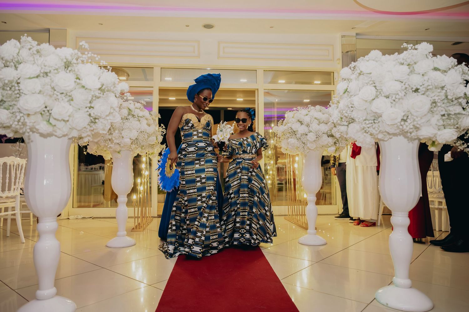 Two women in matching African kente print dresses pose on red carpet at elegant wedding venue.