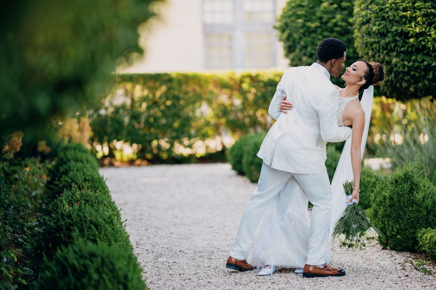 Groom dips bride for a kiss on a gravel garden path surrounded by lush green hedges.