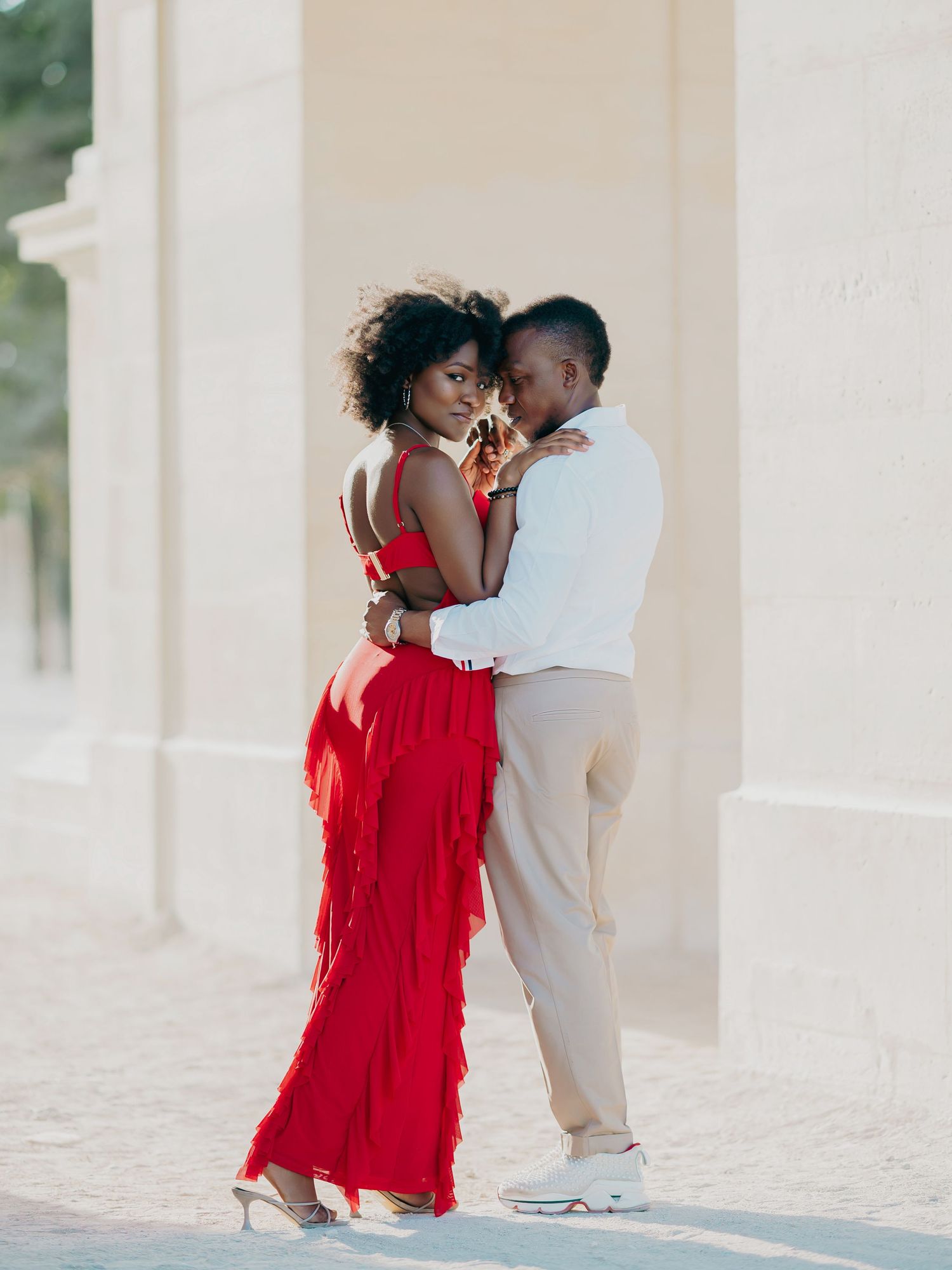 Black couple embracing tenderly against a stone column, woman in dramatic red fringe backless gown and man in white.