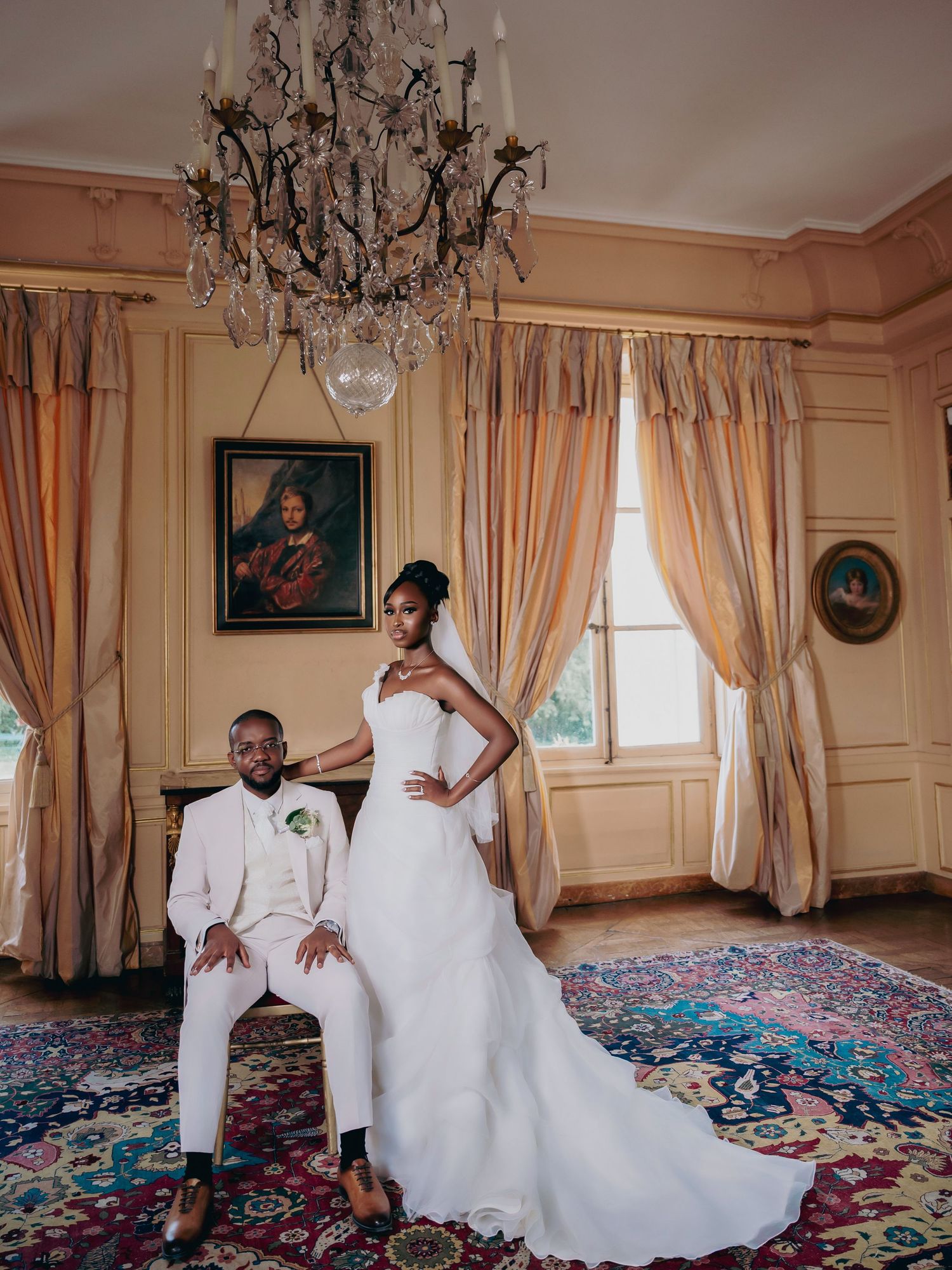 Elegant bride and groom posing in ornate chateau room with crystal chandelier and colorful drapes.