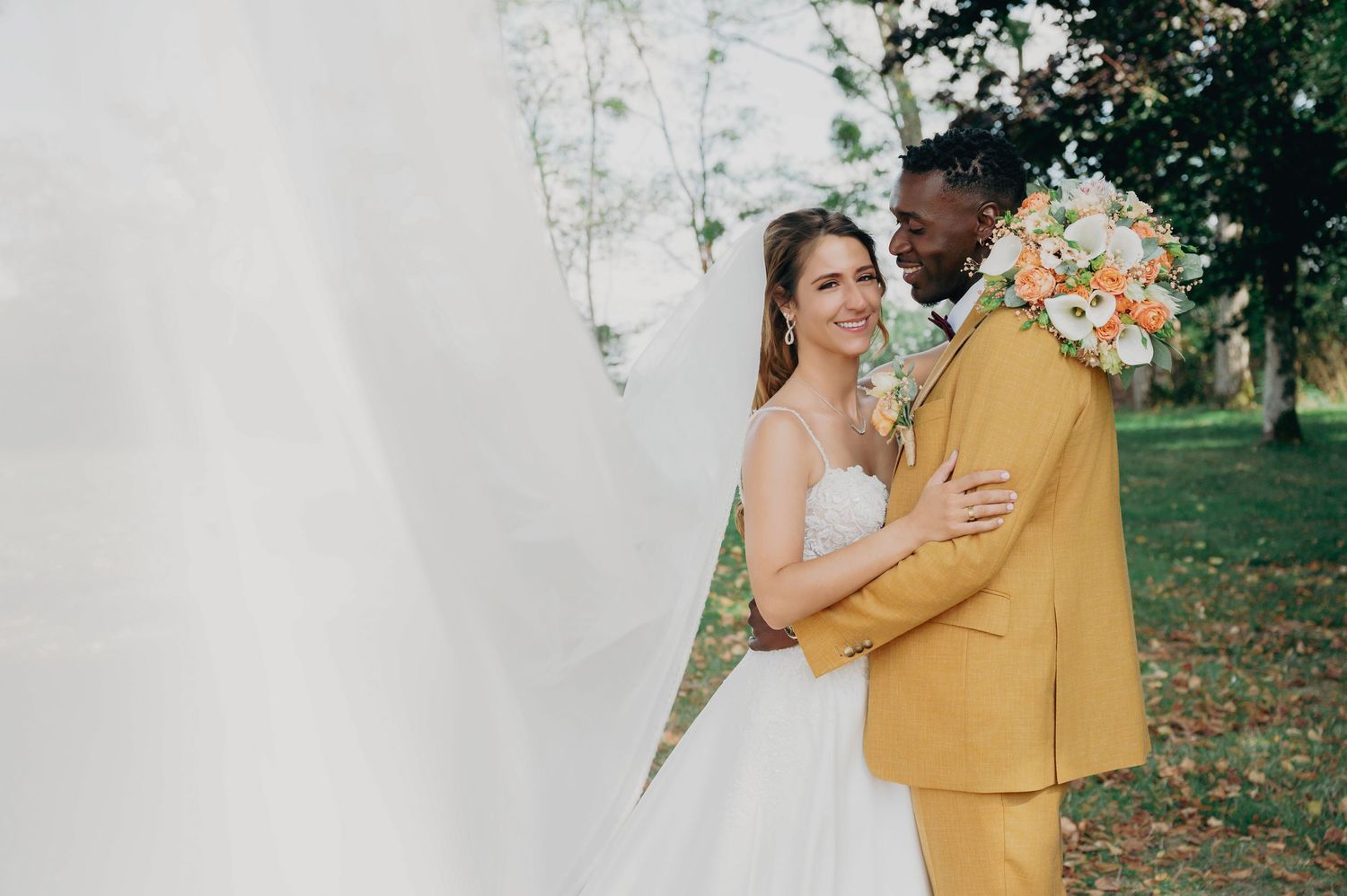 Smiling bride in strapless gown embraces groom in mustard suit holding peach floral bouquet.