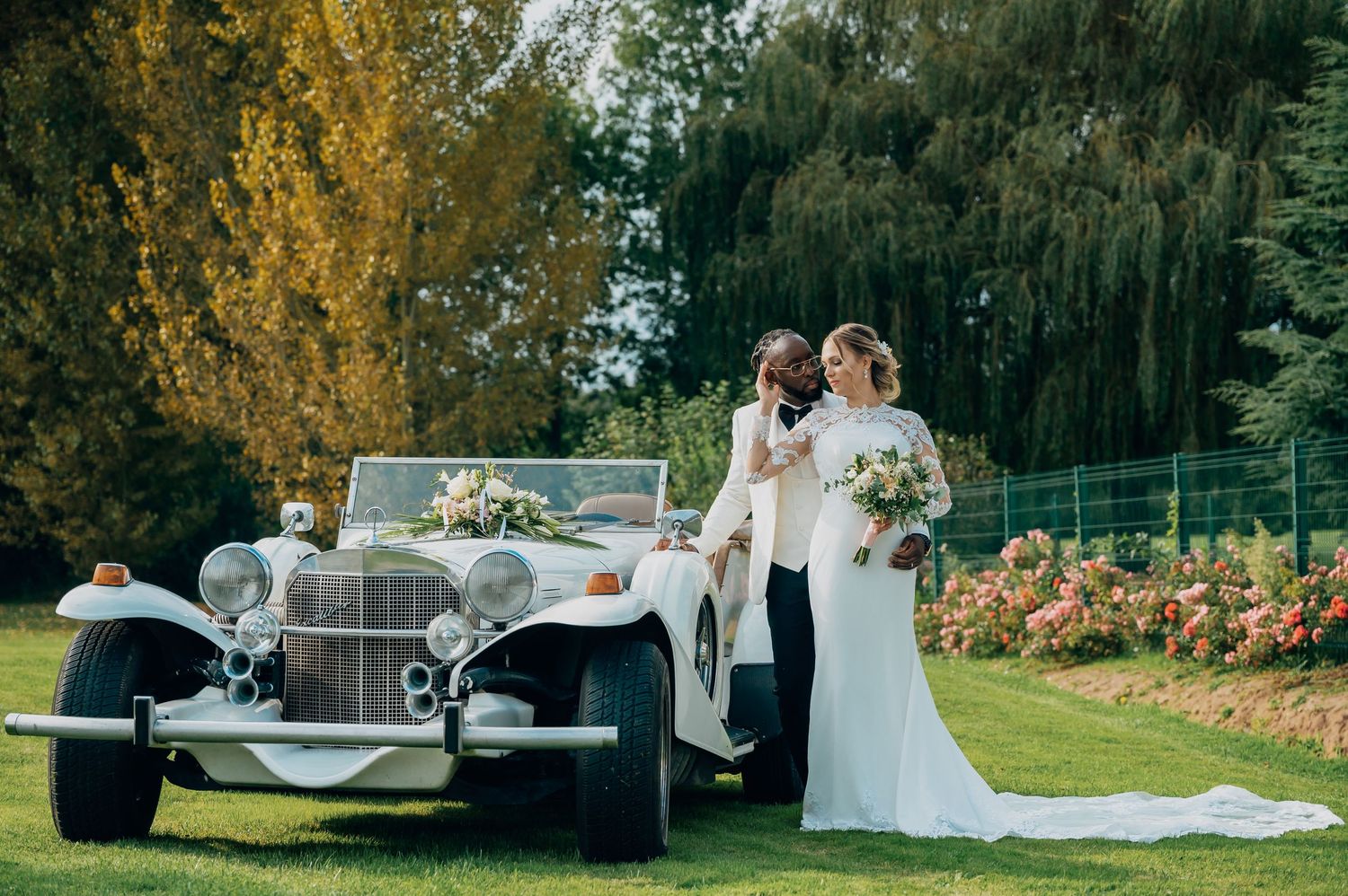 Bride and groom posing beside a vintage white classic car decorated with flowers on a lush green lawn.