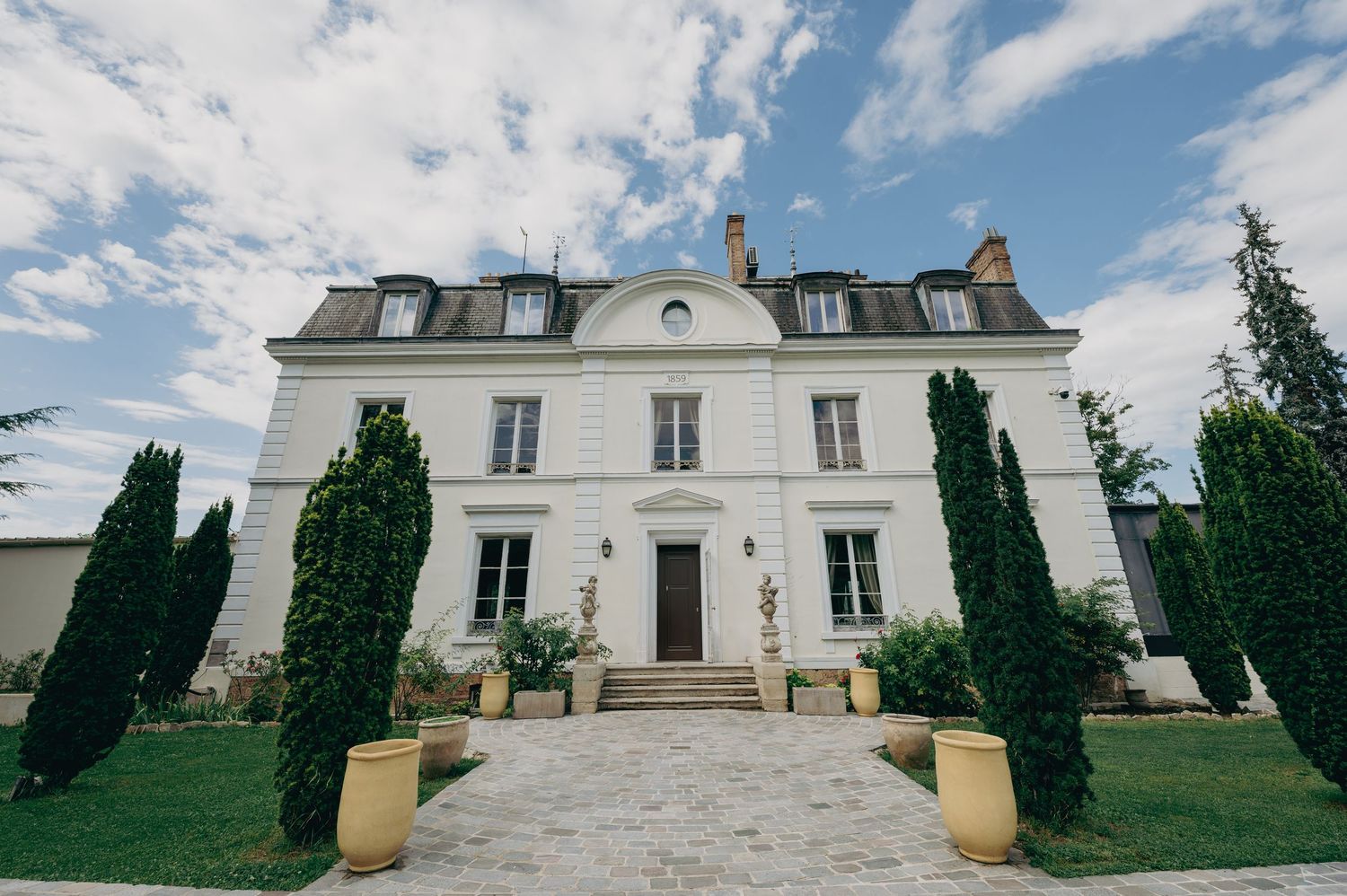 Grand white French chateau wedding venue with manicured cypress trees and gravel driveway under blue sky.